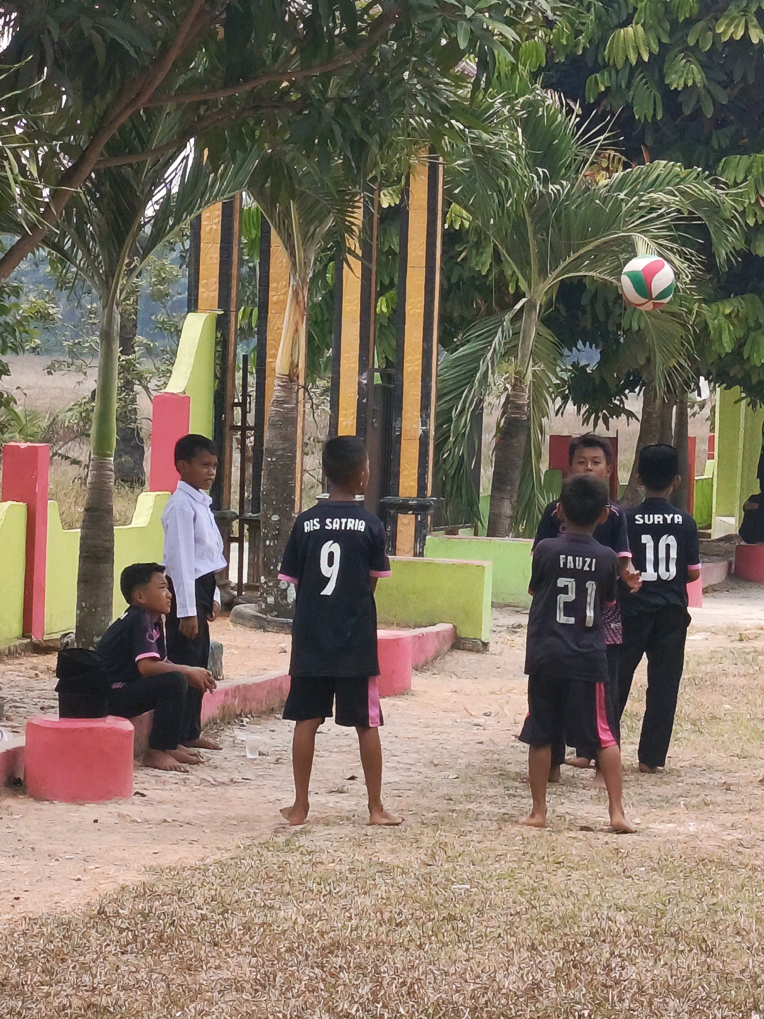 The image depicts a group of young boys in an outdoor setting, possibly a schoolyard or park, in Megang Sakti V, Indonesia. Several boys are wearing dark t-shirts with names and numbers on the back, suggesting they are part of a sports team. One boy in the foreground is wearing a black t-shirt with "FAUZI 21" printed in white. To his right, another boy wears a similar shirt with "SURYA 10." Further left, a boy in a white collared shirt and black pants stands, while another boy in a black t-shirt with "AIS SATRIA 9" is also visible. A volleyball, partially obscured by palm fronds, is suspended in the air towards the upper right of the frame. The environment features palm trees, a dirt ground scattered with dry grass, and brightly colored structures, likely part of a fence or playground equipment, with pink, yellow, and green accents. The lighting suggests it is daytime, with shadows indicating a moderately sunny sky. Some of the boys are barefoot, and one is sitting on a pink circular bench.