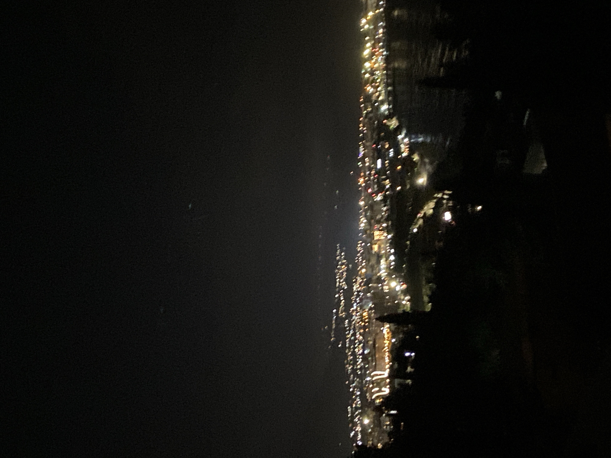The image depicts a nighttime view of Šušanj, Montenegro, from an elevated perspective. The city lights form a bright, linear pattern along the coast. The sea is visible on the right side, reflecting some of the city lights. A dark land mass or vegetation is present on the right side, obscuring part of the city view. The sky is dark and relatively clear, with a few faint lights visible. The overall impression is that of a calm night in a coastal urban environment.