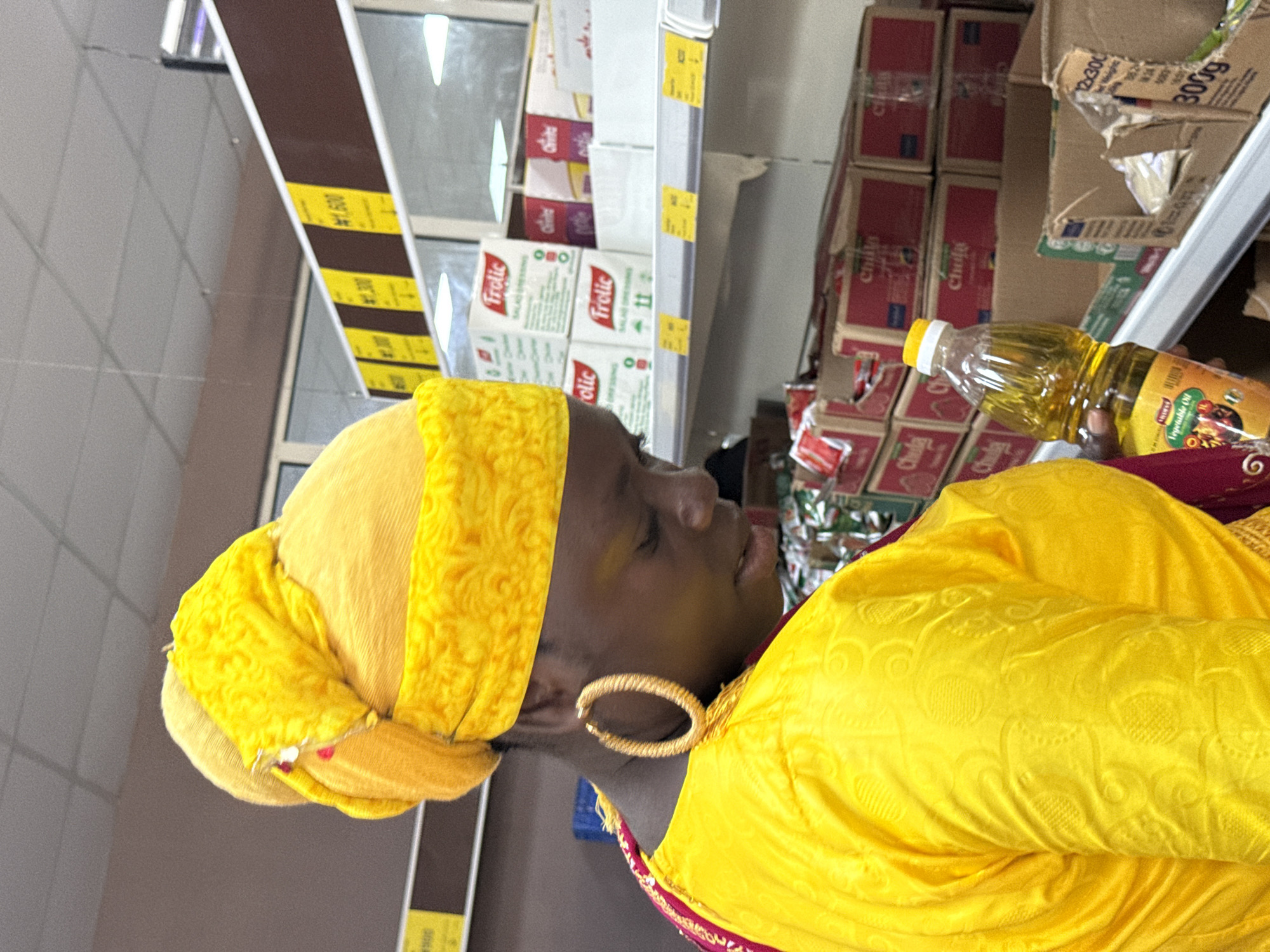 A woman wearing a yellow headwrap and yellow garment stands in a supermarket aisle. She holds a bottle of "Chula Vegetable Oil" in her right hand. Shelves behind her are stocked with food items including boxes of "Frolic Salad Dressing" and what appears to be packages of "Chula" (possibly another food product). Price tags are visible on the shelf edges. The location is likely a supermarket in Iranla, Nigeria, based on the branding on the oil bottle.