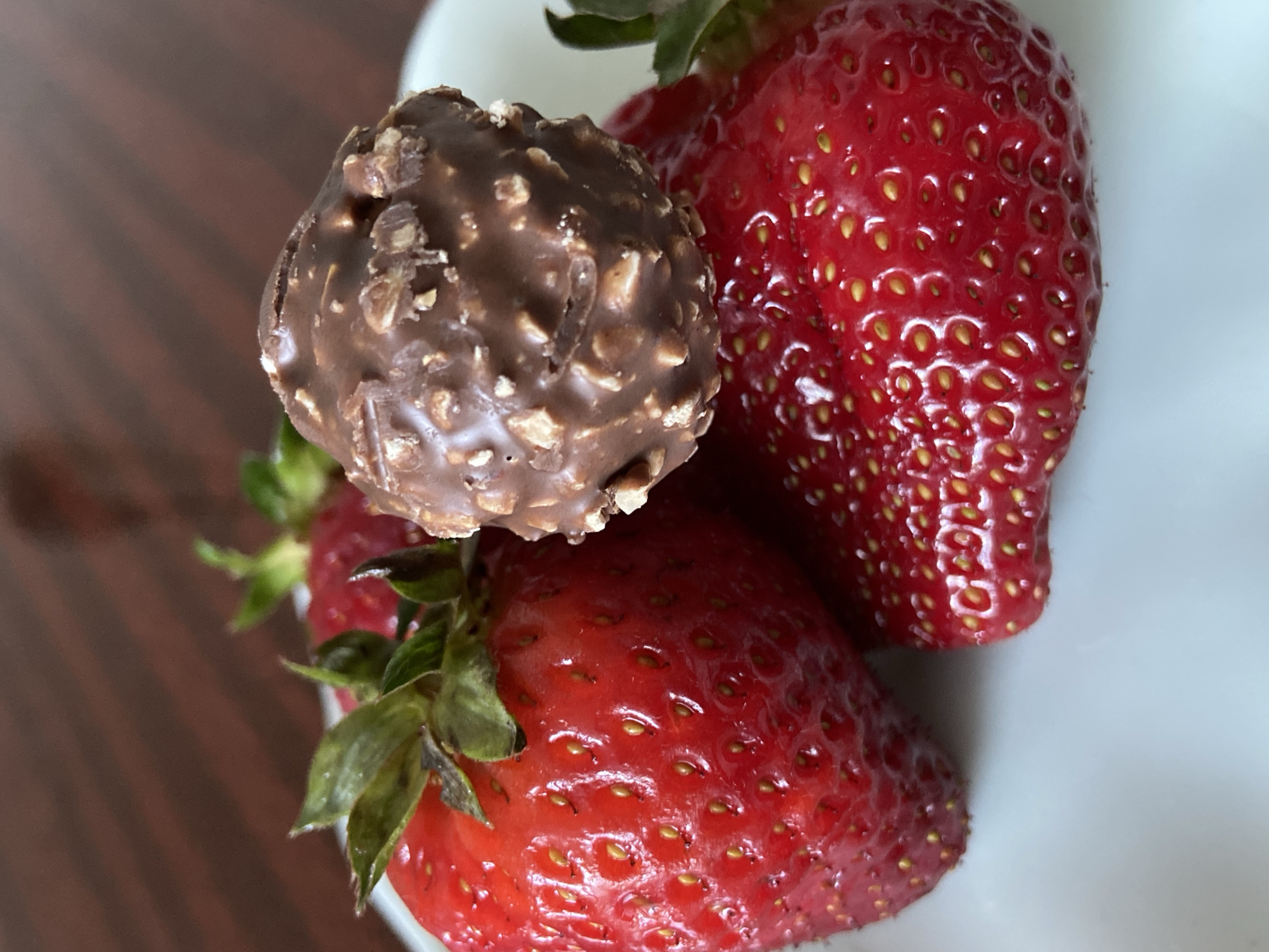 The image displays a close-up, overhead view of a single chocolate confectionery and three fresh red strawberries arranged on a white surface, likely a bowl or plate. A blurred, dark brown wooden surface is visible in the background, suggesting an indoor setting like a table.
The chocolate is spherical and dark brown, covered in numerous small, irregular light-brown pieces, characteristic of chopped nuts or crispy wafer. Its surface appears slightly rough and textured. One prominent crack or indentation is visible on its upper left side.
The three strawberries are a vibrant, glossy red. Their surfaces are dotted with many small, yellow, embedded seeds (achenes), which are typical of strawberries. Two of the strawberries clearly show their green calyxes (leafy tops) and stems, indicating they are fresh. One large strawberry occupies the top right portion of the frame, another is positioned towards the bottom left, and a third is partially visible behind the chocolate and the bottom-left strawberry. The strawberries appear plump and ripe.
The lighting is soft and even, highlighting the textures of both the chocolate and the fruit. The composition creates an inviting visual of a sweet treat alongside fresh fruit. There is no visible text in the image.