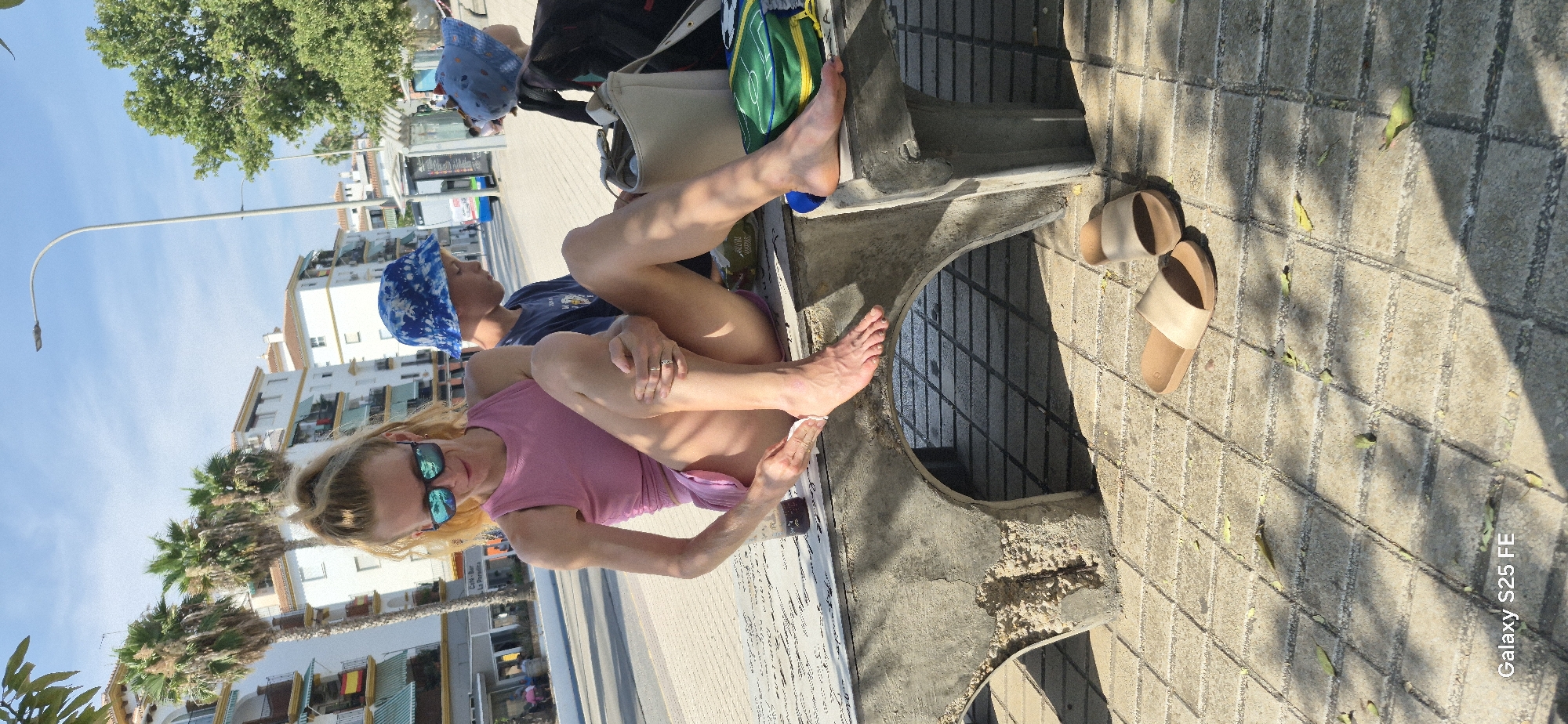 The image depicts a sunny outdoor scene on a paved walkway in Torre Del Mar, Spain. In the foreground, a woman with blonde hair pulled back, wearing sunglasses and a pink tank top with a matching skirt, is seated on a concrete bench. She is attending to her right foot, holding a small white object, possibly a tissue or wipe, and a dark bottle. Beside her, a young boy wearing a blue bucket hat with a white pattern and a dark blue t-shirt is seated, looking away from the camera. To the right of the boy, another young person wearing a similar blue bucket hat is partially visible, along with a black backpack and a light-colored bag. A pair of beige slides is placed on the tiled ground in front of the bench. In the background, white buildings with balconies and palm trees are visible, along with a lamppost and a street. The ground is paved with square tiles, and shadows from trees and an overhead structure are cast across it.