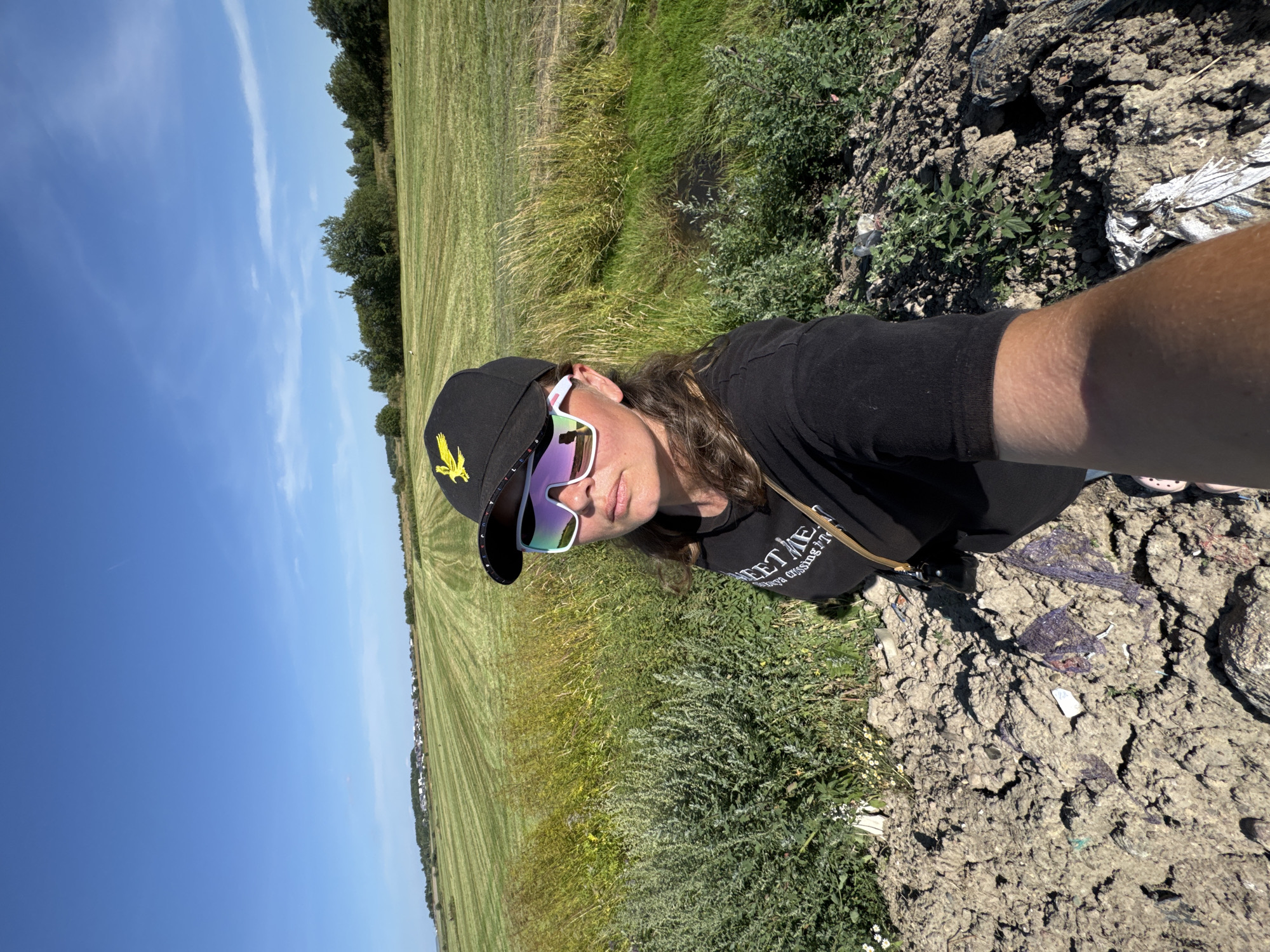 In Kempelevo, Russia, a woman takes a selfie in a rural setting under a clear blue sky. She wears a black baseball cap with a yellow eagle emblem and oversized white-framed sunglasses. Her black t-shirt has white text that reads "SWEET ME" and "ya cossing T". She stands in a patch of rough, dry ground with some sparse vegetation. Behind her, a mowed field stretches out, dotted with trees along the horizon. A small cluster of buildings is visible in the distance. The overall scene suggests a sunny, outdoor recreational or observational moment in a countryside area.