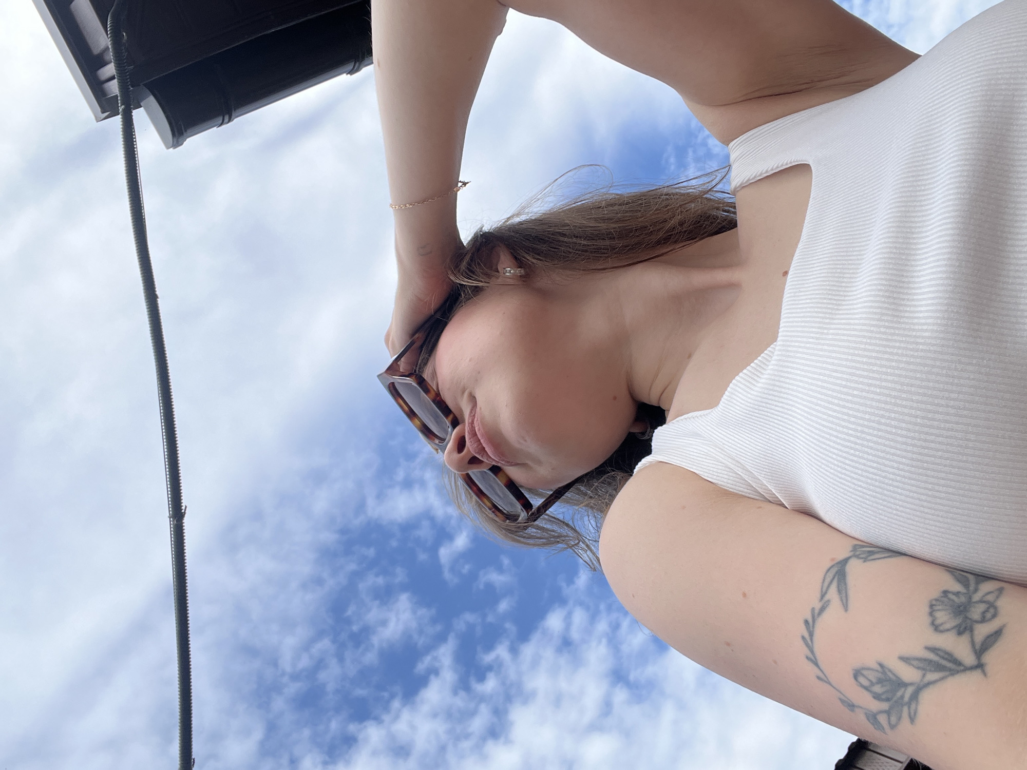 A low-angle, upward-facing outdoor photograph captures a young adult female from the chest up, set against a blue sky with scattered white clouds in Chaikovsky, Russia. The subject's head is tilted back, and her face is turned upwards and slightly away from the viewer, with her eyes obscured by tortoiseshell sunglasses. Her right hand (viewer's left) is raised, with fingers touching the side of her head and hair, possibly adjusting the sunglasses or shielding her eyes. A delicate gold-toned chain bracelet with a small charm is visible on her right wrist, and a small silver-toned hoop earring is in her right ear. She wears a white, ribbed tank top with vertical stripes and square-cut straps. A faded grey tattoo, depicting a circular floral and leaf wreath, is visible on her upper right arm. In the upper left quadrant of the frame, a dark, corrugated flexible pipe or cable extends horizontally. This cable appears to connect to a dark brown/black building structure, likely a roofline and gutter system, visible in the upper right quadrant.