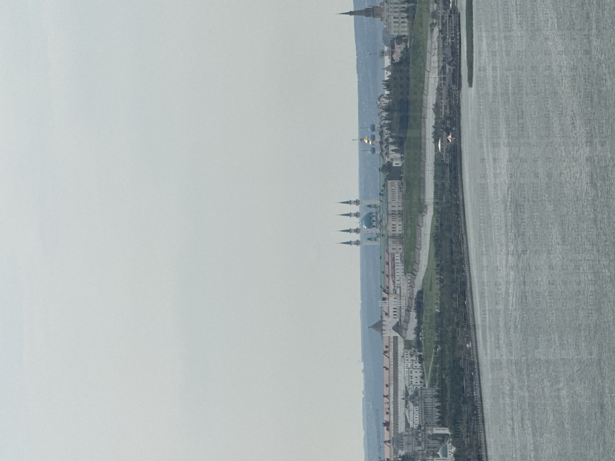 This image shows a panoramic view of the Kazan Kremlin, a historic fortified complex located in Kazan, Russia. The Kremlin is situated on a hill overlooking the Kazanka River.
In the foreground, the wide, grey expanse of the Kazanka River dominates the lower half of the image. The water appears calm with subtle ripples.
The middle ground displays the complex of buildings that comprise the Kazan Kremlin. Prominent structures include the Kul Sharif Mosque, recognizable by its blue domes and white minarets, and the Annunciation Cathedral, characterized by its gold domes. Other buildings within the Kremlin complex, featuring white walls and red-tiled roofs, are also visible. Trees and green spaces are interspersed among the buildings. A wall and pathways are also discernible within the Kremlin grounds.
In the background, the horizon is marked by a faint, blue-tinged landmass, possibly hills or the far bank of the river. The sky is overcast, appearing a uniform, pale grey, suggesting a cloudy day. The overall atmosphere is serene and historical. No people are clearly discernible in the image.