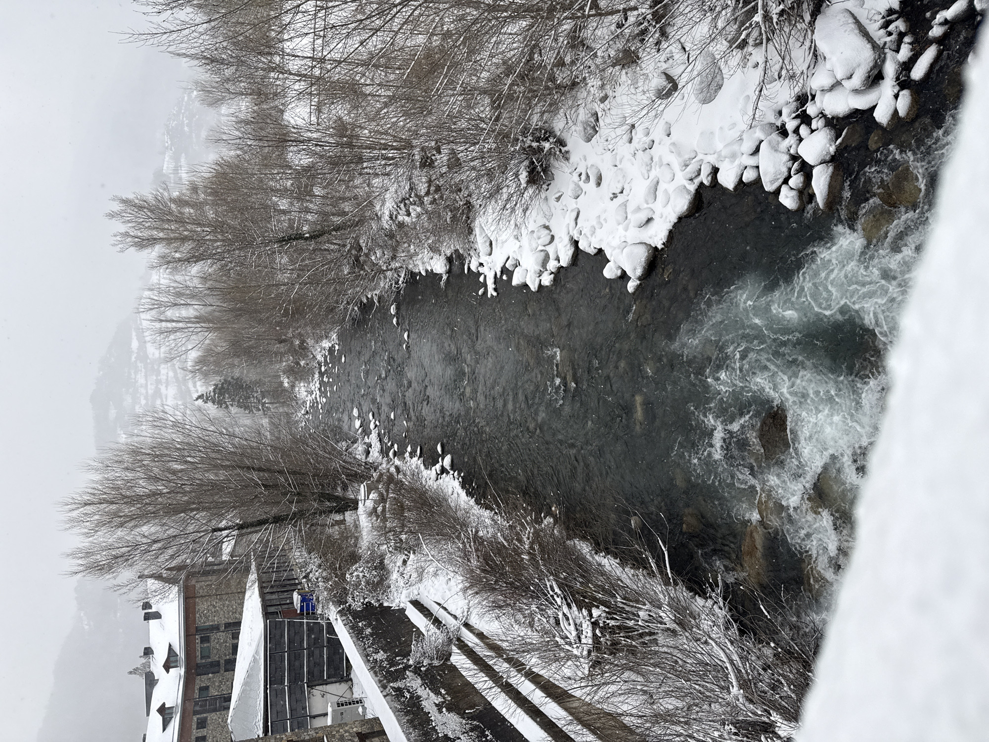 The image depicts a river flowing through the town of Benasque, Spain, during a wintery season. The riverbank is covered in snow, which also clings to the rocks along the edges. Bare trees and bushes line the riverbanks, dusted with snow. A building is seen to the left. The water appears to be flowing rapidly, with some whitewater sections. A bridge or walkway appears in the foreground.