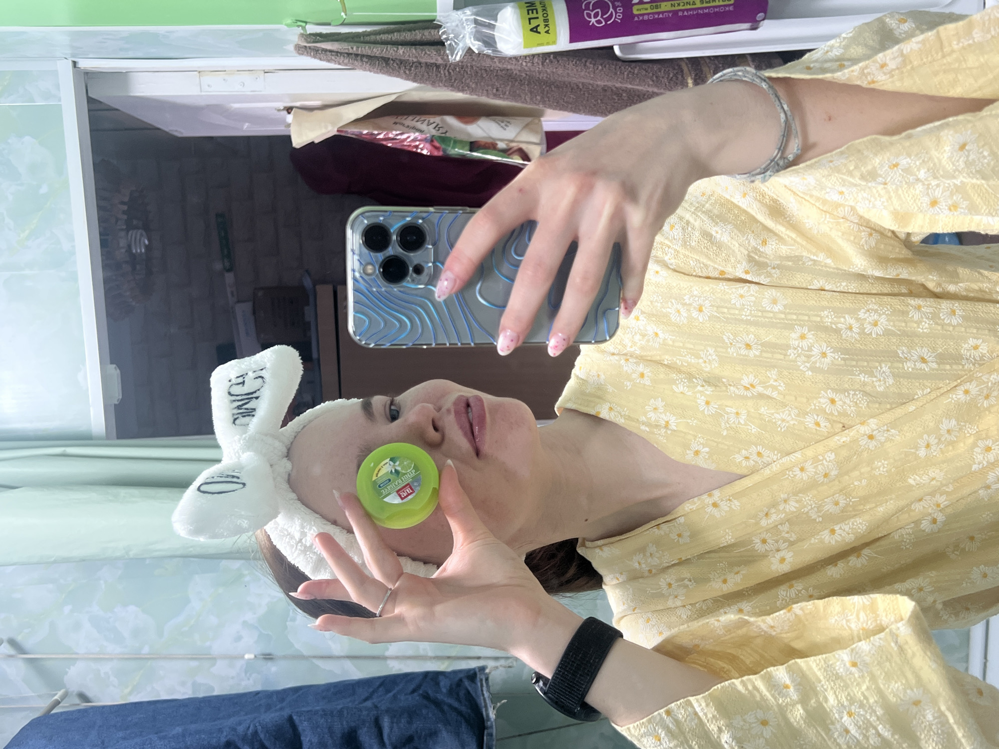 A woman in a yellow floral robe and a white bunny-ear headband is taking a selfie in a bathroom. She is holding a green jar of facial cream in her left hand. She is using a phone with a light blue patterned case. The bathroom has green and white tiles. On a shelf above the woman, there are several items including cotton pads. The exact location within the city of Chaikovsky, Russia cannot be determined from this image.