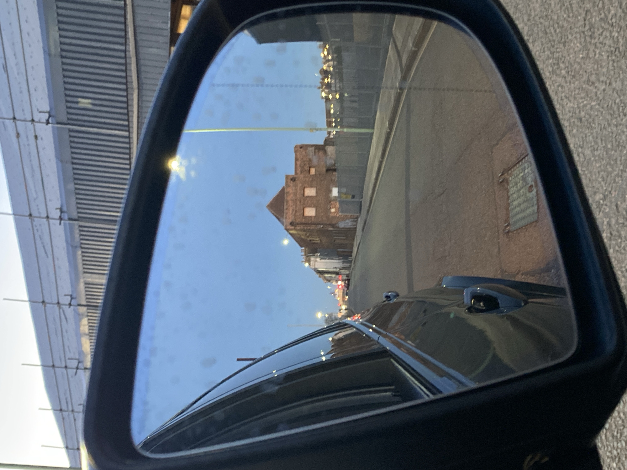 The image shows a car's side mirror reflecting a street scene in East Suffolk, United Kingdom. The reflection includes buildings, a street, a lamppost, and other vehicles. The building with a visible roof, likely located at 1-9 Grimwade Street, Ipswich, United Kingdom is reflected through the rearview mirror. Scaffolding is visible on the left side of the mirror.