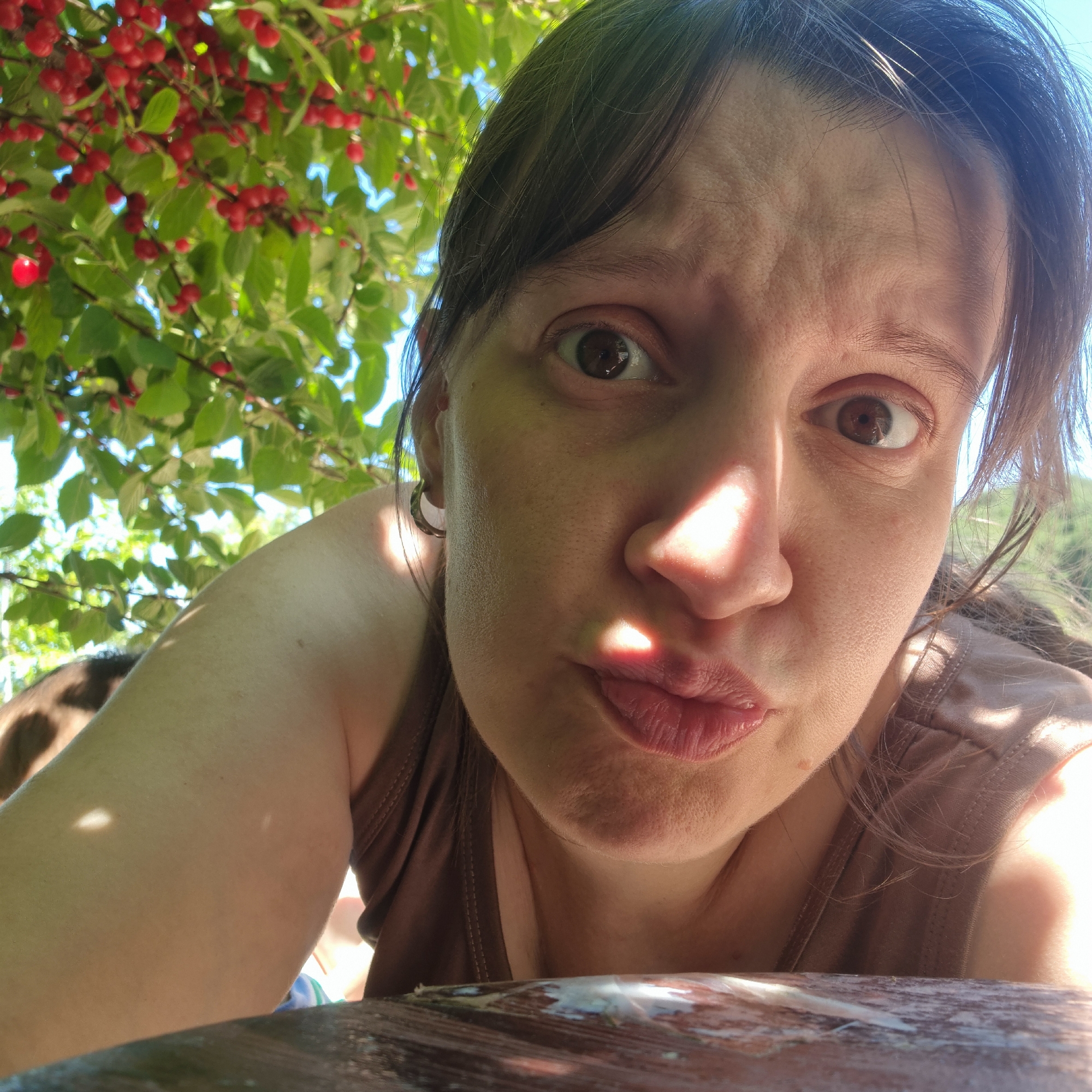 A woman is captured in a close-up, low-angle selfie outdoors, with her face filling most of the frame. She has brown hair and is wearing a brown tank top. Her expression is one of mild surprise or questioning, with her eyebrows raised and her lips pursed. Sunlight highlights her face, casting shadows. In the background, lush green leaves and red berries suggest she is under a tree. The lower part of the image shows a wooden surface, possibly a table.