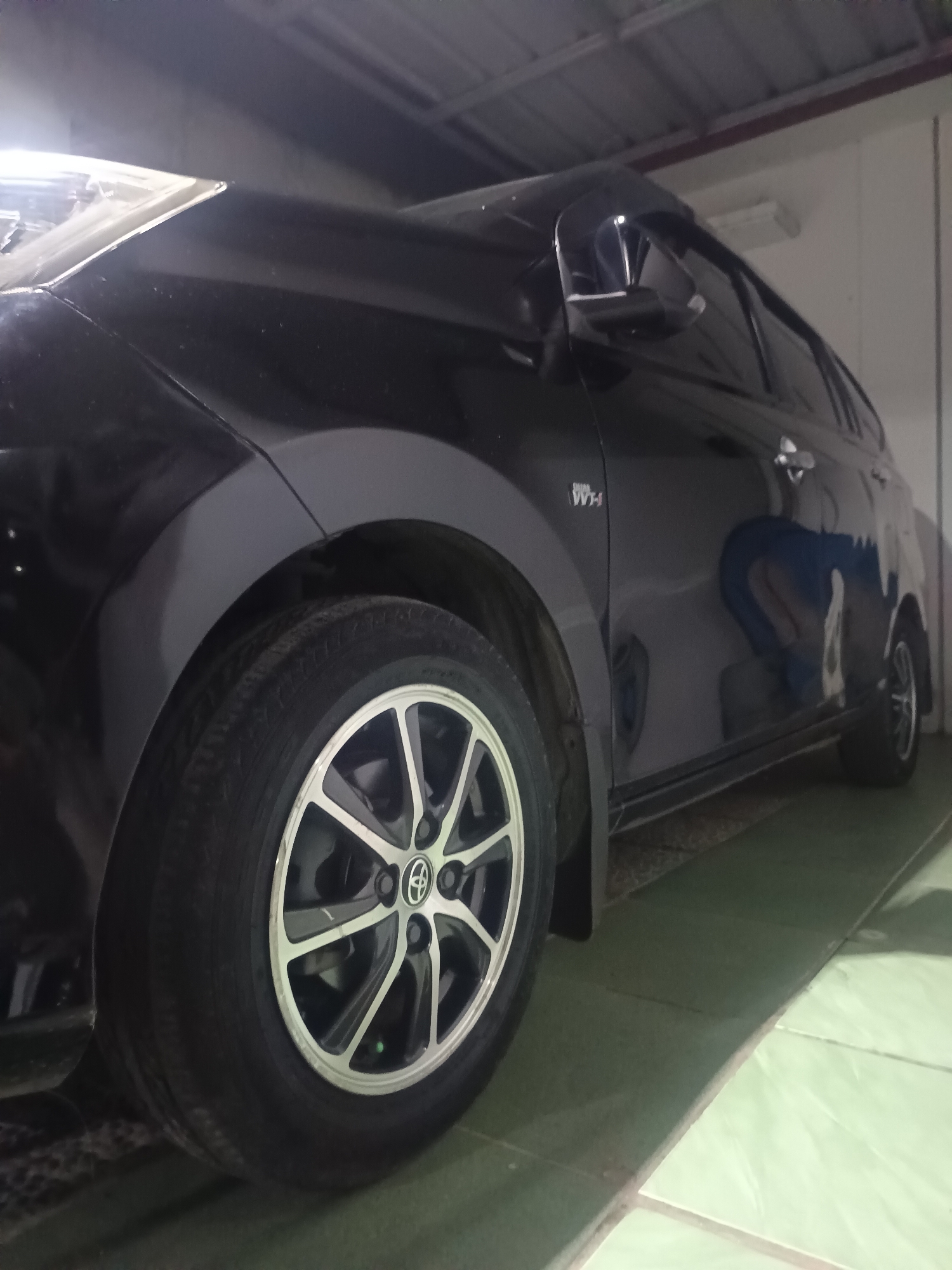 A black four-door vehicle, identifiable by the "Dual VVT-i" emblem on its front left fender and a Toyota logo on the front left wheel hub, is parked indoors on a light green tiled floor in Maleber, Indonesia. The car is oriented with its left side facing the viewer, captured from a low perspective. The front left headlight area reflects significant light. The car is equipped with black tires and multi-spoke alloy wheels featuring a silver and black design; a green valve stem cap is present on the front left wheel. A discernible reflection of a person, bent over, is visible on the car's dark side door panel. The immediate surroundings include a light green tiled floor with a diagonal grout line, and an industrial-style ceiling with exposed beams and a fluorescent light fixture in the upper right background.