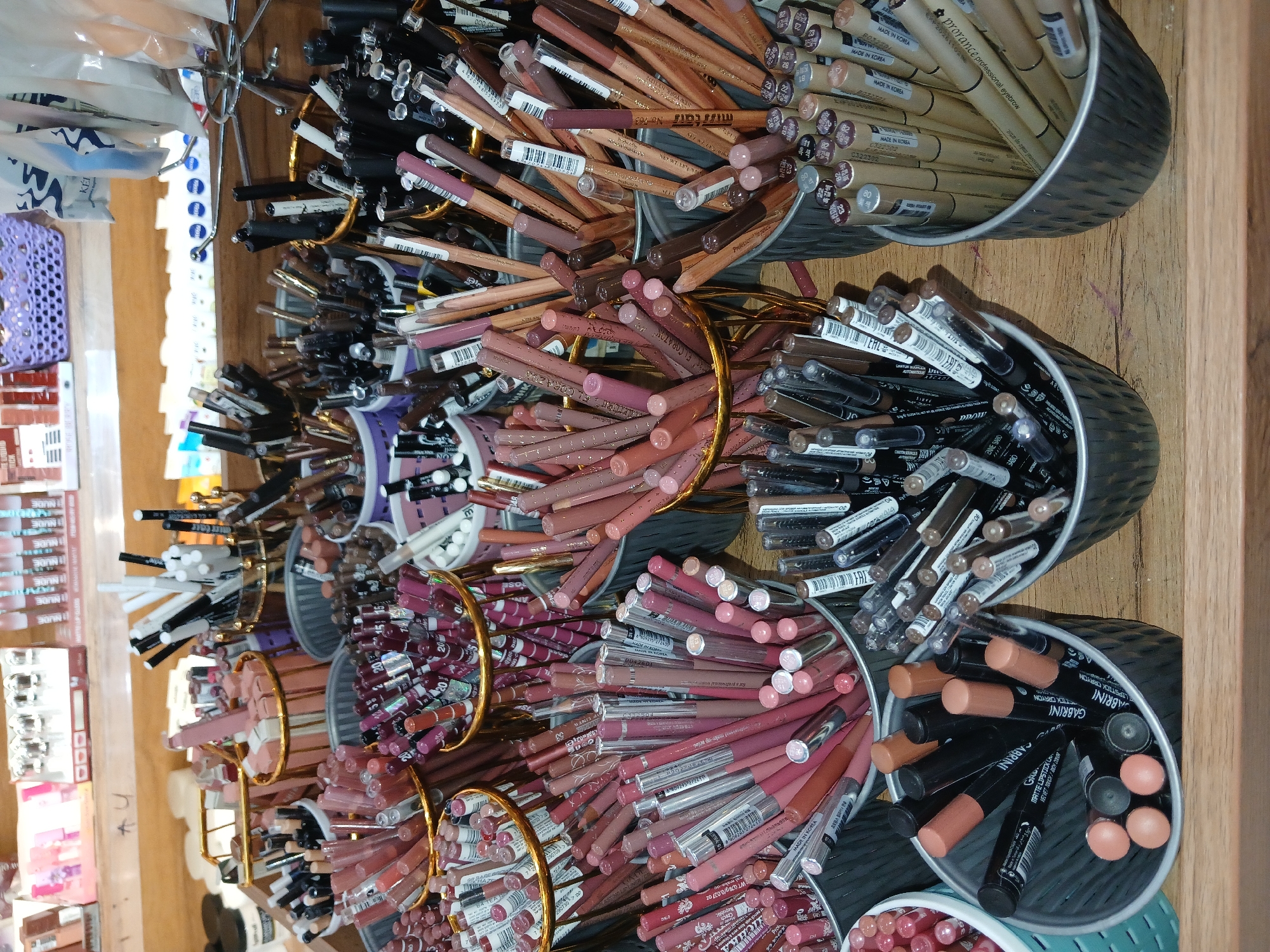 The image displays a close-up, eye-level view of a retail display of makeup, specifically lip liners, eyeliners, and eyebrow pencils, arranged in various buckets and containers on a wooden surface. The items are predominantly in shades of pink, nude, brown, and black, with some metallic accents. The products are neatly organized and appear to be new and unused. The background shows shelves stocked with more cosmetic products and some woven baskets, suggesting a retail environment. The lighting is bright and even, indicating it's likely an indoor setting. The scene is static, with no people or immediate actions depicted. The image is likely taken in a makeup store or a cosmetics section of a larger retail establishment. Specifically, this display is located within the "Cosmopolitan" makeup store at the MEGA PLANET shopping mall in Tashkent, Uzbekistan.