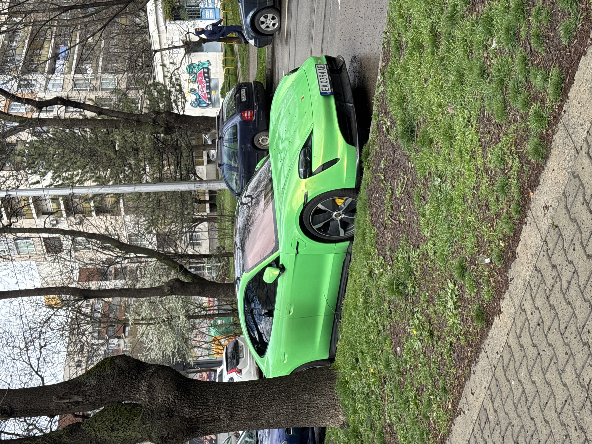 A green car is parked next to a sidewalk and a grassy area, with a tree in the foreground. Other cars are parked behind it. A person is seen standing near a building with graffiti. The location appears to be urban, with apartment buildings visible in the background. The city is unknown.