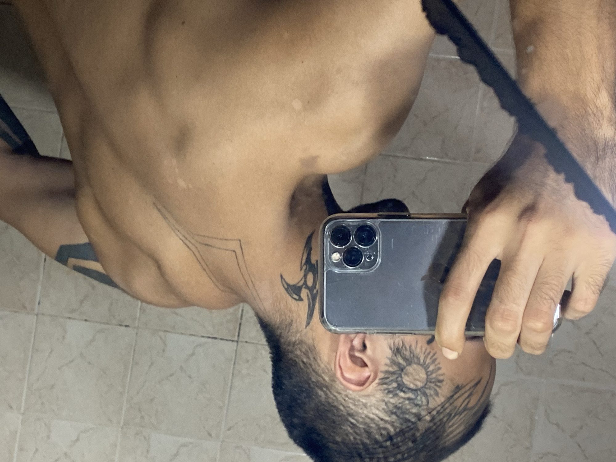 A shirtless man with tattoos is taking a selfie in a bathroom with tiled walls. He holds a smartphone in his right hand, partially obscuring his face. Tattoos are visible on his head, neck, chest, and arms.