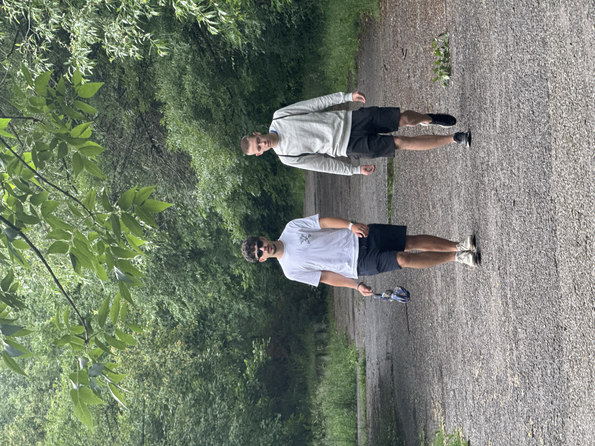 The image captures two young men standing on a paved path, surrounded by dense, lush green foliage in what appears to be a park or natural area in Sofia, Bulgaria. The path, made of grey, gravel-like material, extends into the background, framed by abundant trees and bushes that create a verdant, tunnel-like environment.
In the foreground, positioned slightly to the right of the path, a man with dark, curly hair and sunglasses is visible. He wears a white t-shirt featuring black text that reads "THE NORTH FACE" (vertically arranged with a logo), dark shorts, and light-colored sneakers. A watch adorns his left wrist, and he holds a blue-patterned item, possibly an umbrella or a small bag, in his right hand.
Further along the path, to the left, stands a second man with short, light-colored hair. He is dressed in a light grey long-sleeved top, revealing a blue collar underneath, dark shorts, and dark sneakers. A strap, possibly from a bag, crosses his shoulder.
Both men are standing and looking towards the camera with neutral expressions, suggesting they are either posing for a photograph or taking a break from a walk. The natural lighting indicates it is daytime, likely a mild or overcast day given the soft illumination, which is consistent with spring or summer due to the vibrant green vegetation. The scene conveys a tranquil, outdoor setting.