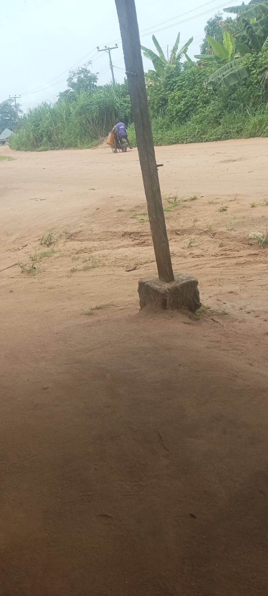 A man is riding a motorcycle on a dirt road in Amauzu, Nigeria. The road is flanked by dense vegetation. An upright wooden post is embedded in a square concrete base near the foreground. Overheard power lines are visible in the background.