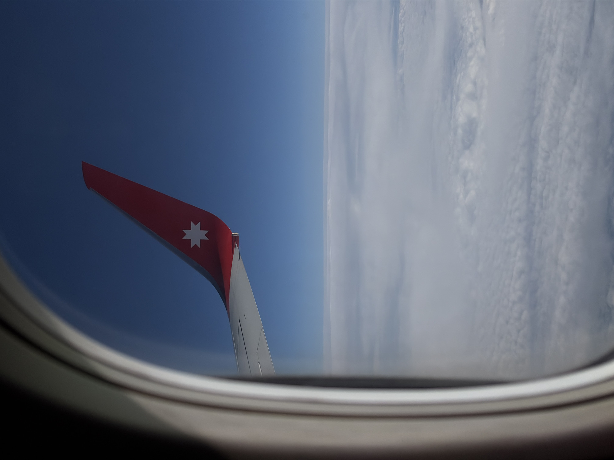 The image depicts an airplane wing visible through a circular airplane window. The wing is red with a white star emblem and extends into a bright blue sky on one side and a dense cloud formation on the other. The window frame is white. No people are visible. The location cannot be definitively determined as Domodedovo, Russia, based solely on the visual information.