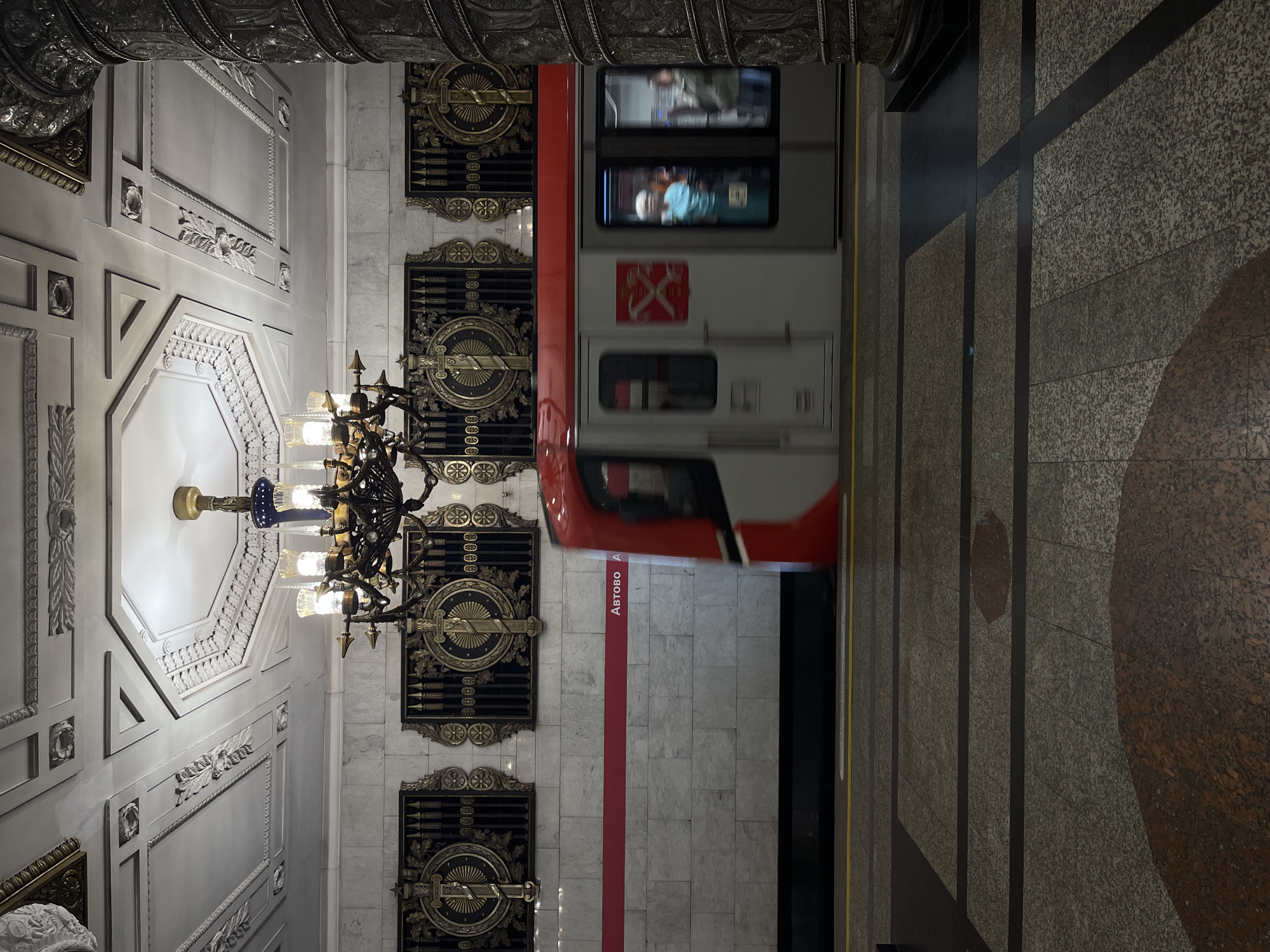 This is a photograph of the Avtovo metro station in Saint Petersburg, Russia. The image captures a metro train in motion, its red and white carriage blurred as it passes by the platform. The station's interior is grand, featuring a highly ornate ceiling with intricate plasterwork and a large, multi-tiered chandelier. The walls are clad in marble and adorned with decorative bronze panels. A red stripe runs along the wall above the platform, with the station name "АВТОВО" (Avtovo) visible. The platform floor is a mosaic of granite tiles in various shades of gray and brown, with black and yellow lines indicating the platform edge. In the windows of the train, blurry figures of passengers can be seen. A large, ornate column, possibly covered in decorative metalwork, stands on the right side of the frame.