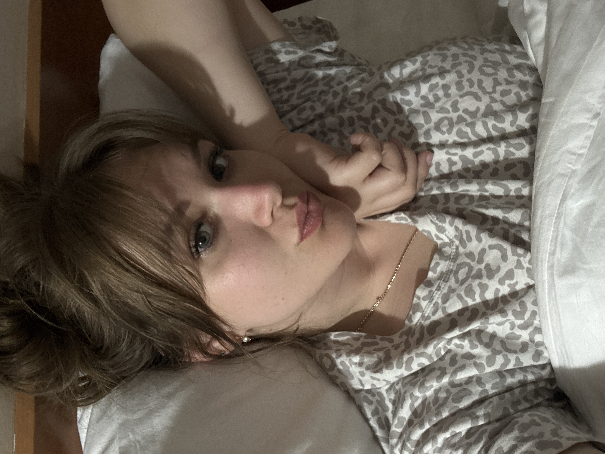 A woman lies in a bed with white sheets and a gray and white leopard-print top. Her head rests on a white pillow. She has fair skin, light brown hair, and blue eyes. Her left arm is raised, with her hand near her face. She is wearing a delicate gold necklace and a stud earring. The scene appears to be indoors, possibly a bedroom. There is no indication in the image that allows for the determination of the location in the city of Yevpatoriya, Ukraine.