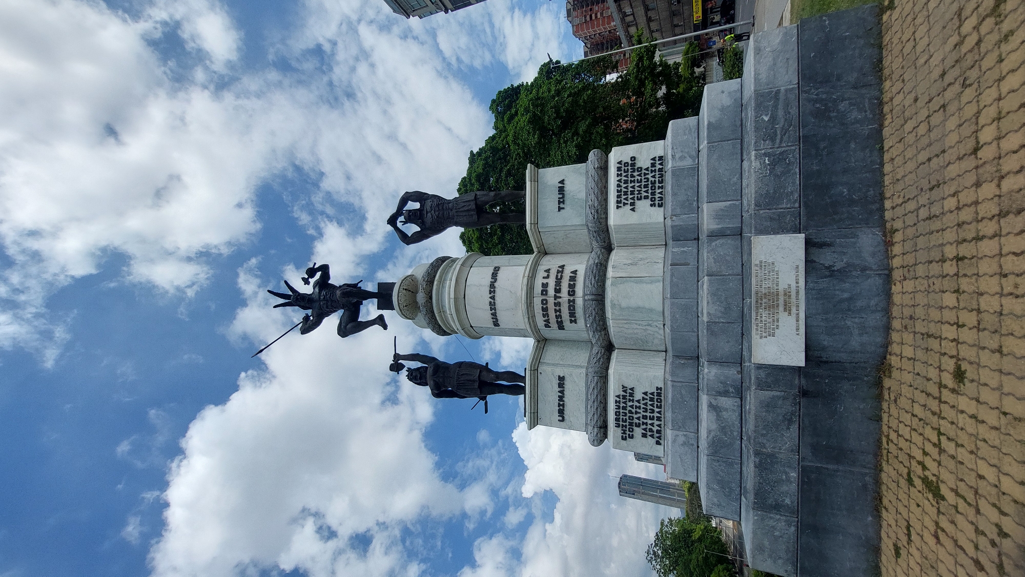 The image depicts the "Paseo de la Resistencia Indígena" monument located in Caracas, Venezuela. Three indigenous figures stand on the monument: Urimare, Tiuna, and Guaicaipuro, each with accompanying plaques. The central pillar of the monument has "Paseo de la Resistencia Indígena" engraved on it. A separate plaque at the base of the monument contains an inscription. The monument is constructed of grey stone, with a multi-tiered base. The environment consists of a partly cloudy sky, a paved ground, trees, and nearby buildings.
