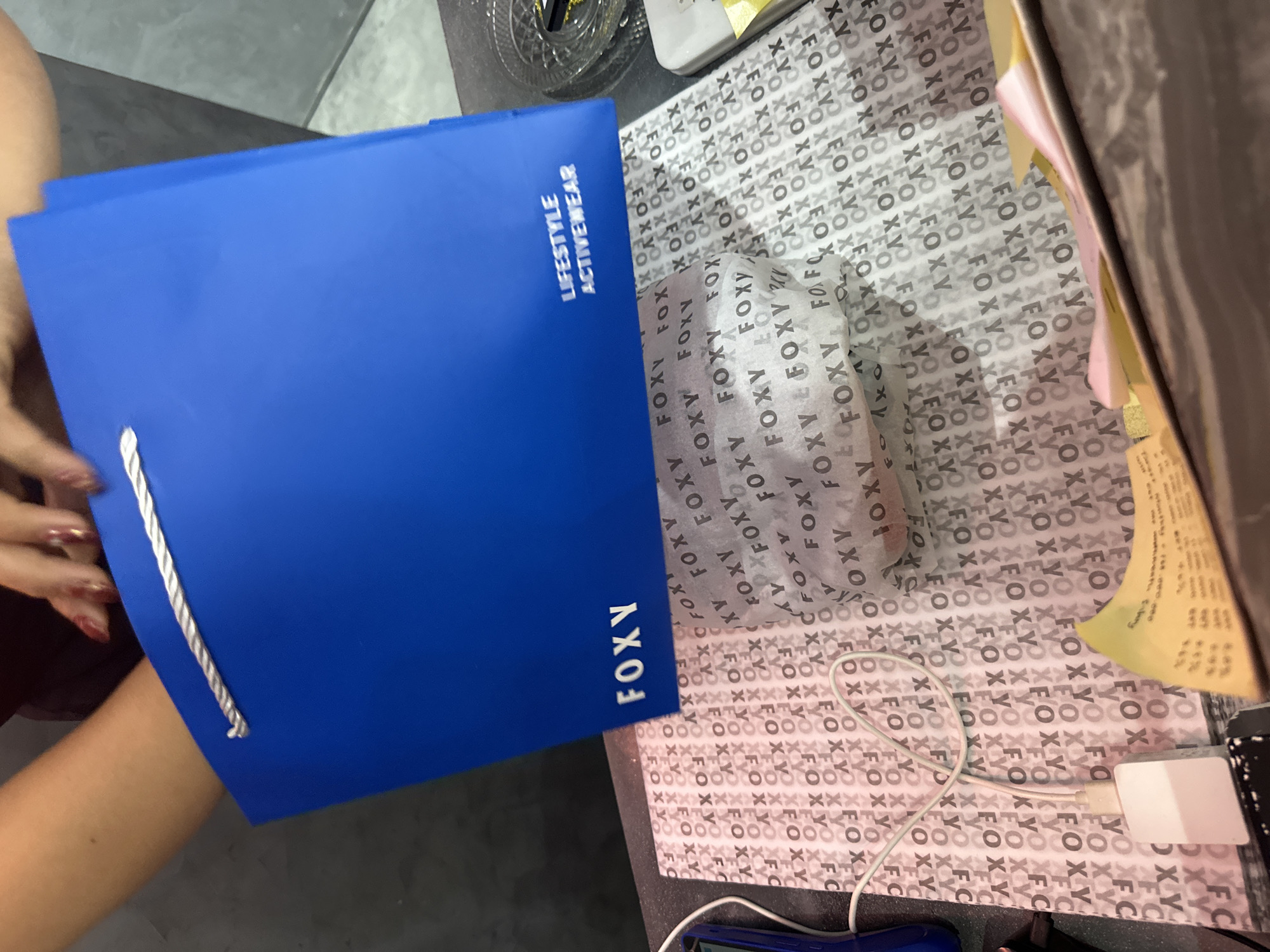 In this close-up, eye-level indoor shot, a person's hands, adorned with red-nailed polish, are shown holding the top of a bright blue shopping bag. The bag, with white twisted handles, is open, revealing a glimpse of tissue paper inside. The tissue paper is patterned with the repeated word "FOXY." Below the bag, a charging cable for a mobile device snakes across the surface, plugged into a white adapter. In the foreground, a yellow sticky note with some text is visible. The background is blurred, but appears to be a retail environment. The image does not contain information to determine the location in the city of Ubud District, Indonesia.