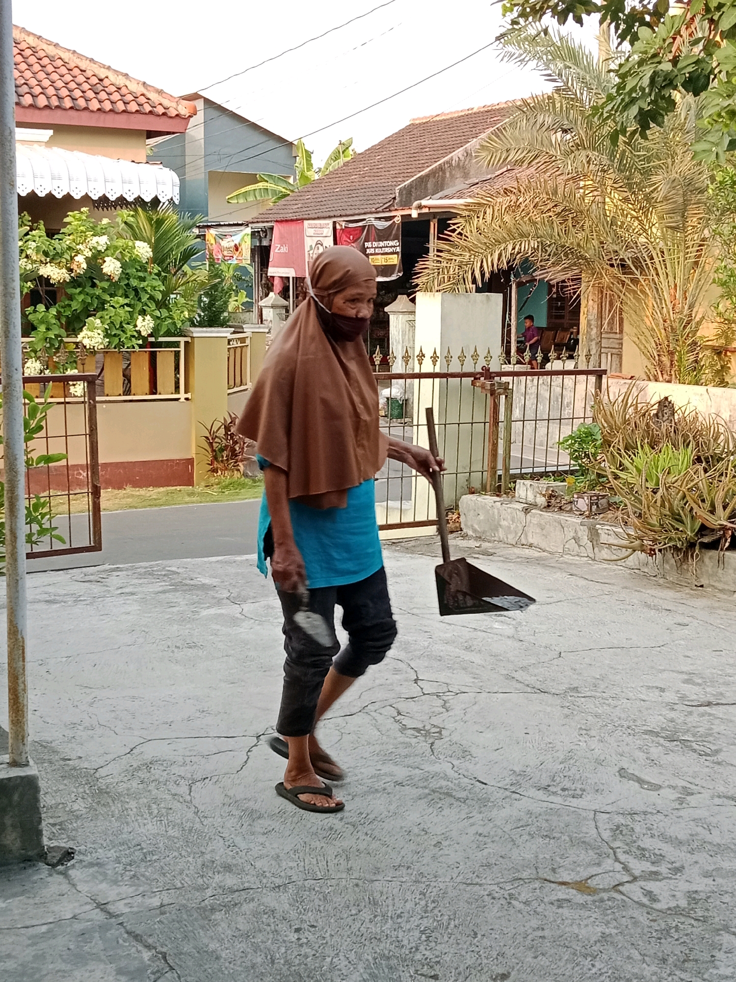 In Waru, Indonesia, an elderly woman wearing a brown hijab and a blue shirt walks on a cracked concrete path, holding a dustpan and a broom. The woman is also wearing a black face mask. In the background, houses with red tile roofs are visible, along with trees and a metal fence. A child can be seen standing near the entrance of one of the houses. The scene appears to be a residential area during daylight.