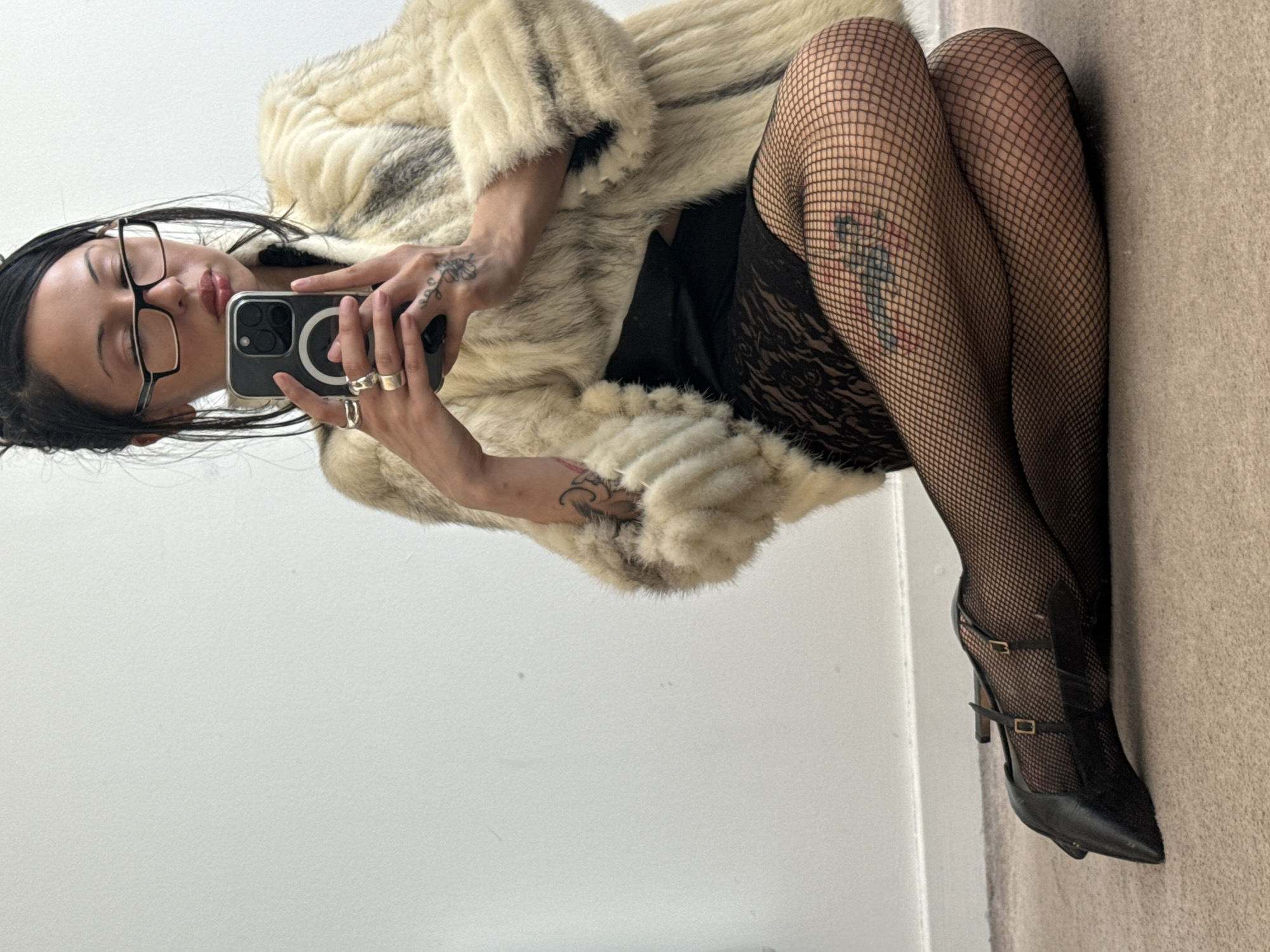This rotated image captures a person taking a mirror selfie indoors, likely in London, United Kingdom. The subject, with dark hair and wearing black-framed glasses, is seen looking towards their smartphone, which they hold in their right hand. The phone, an iPhone model with multiple cameras and a visible MagSafe ring, is angled to reflect their image.
The person is dressed in a plush, light-colored (cream or beige) fur coat. Underneath, they wear black fishnet stockings and black lingerie or shorts with lace details visible on the thigh. Their legs are bent, and their feet are adorned with black high-heeled shoes featuring straps and buckles. Multiple silver rings are visible on their fingers.
Notable details include several tattoos: a delicate floral or vine-like tattoo on the left hand/wrist and a colorful, abstract figure tattoo visible on the right thigh through the fishnet stockings. The background consists of a plain white wall on the left side of the frame and a beige or tan carpet on the right. The image's orientation suggests it was originally captured horizontally or vertically and then rotated 90 degrees counter-clockwise. There is no visible text in the image, and the indoor setting does not provide clues about the time of day.