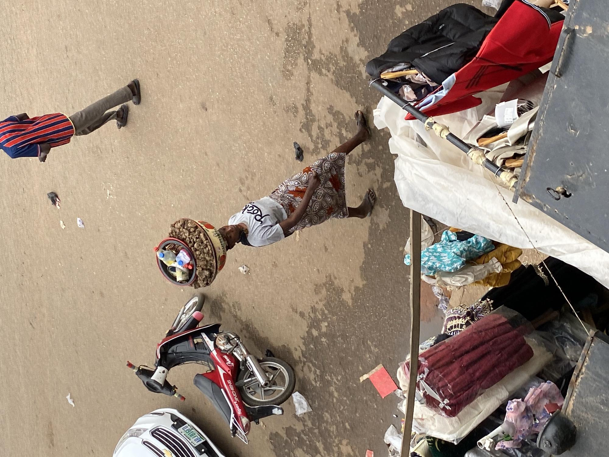 The image depicts a street scene, possibly in Bukuru, Nigeria, judging by the presence of license plates, architectural styles, and clothing.
A woman is seen carrying a large basket-like container on her head. Her basket appears to be filled with various items.
A man is walking on the left of the image. He is wearing gray pants and a striped shirt.
A red motorcycle lies on the ground near a white car.
A container is situated in the background of the image, filled with assorted clothing and objects, possibly for sale.
The ground is concrete.