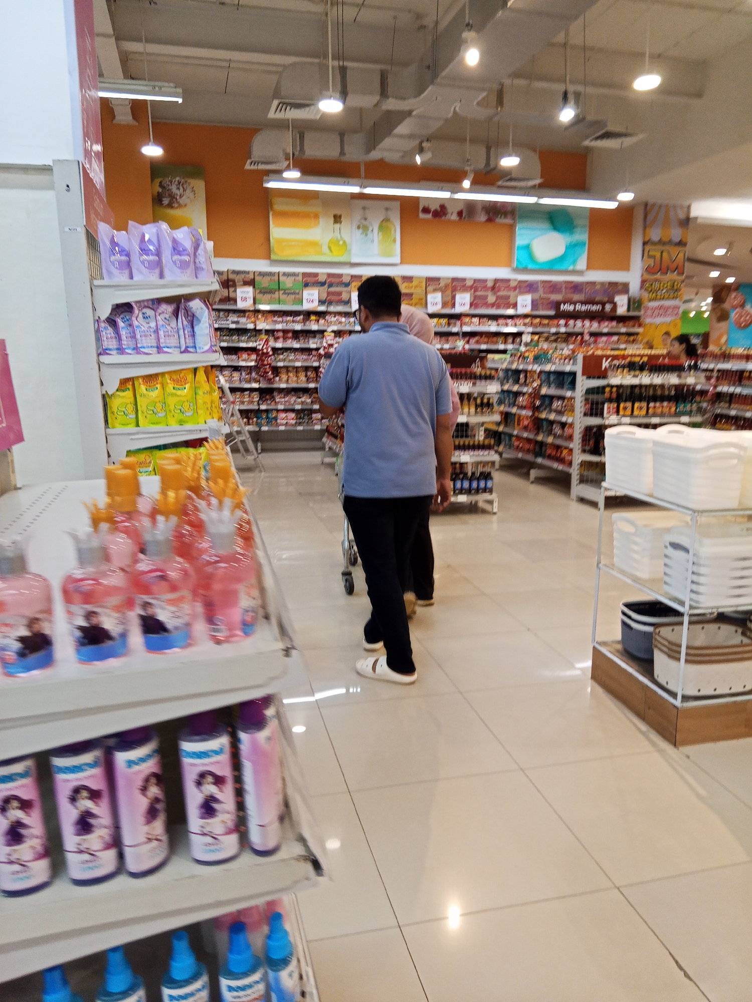 A mid-shot, eye-level, full-color photograph taken from a slightly elevated perspective captures an aisle within a brightly lit supermarket in Lubuklinggau, Indonesia. The scene depicts an interior retail environment with multiple shelves stocked with various products. In the foreground, a shelving unit displays rows of pink and purple liquid soap bottles, some with cartoonish characters on their labels. Another shelf above holds yellow packaged goods. Further down the aisle, a man in a blue short-sleeved shirt and black pants is pushing a shopping cart, walking away from the camera. Another individual, partially visible, walks ahead of him. The supermarket aisle is lined with extensive shelving units filled with a variety of packaged goods, including food items such as instant noodles, indicated by a sign reading "Mie Ramen," and other consumer products. The floor is tiled in a light, neutral color. Overhead lighting fixtures are visible, contributing to the overall bright illumination of the store. The perspective offers a view down the length of the aisle, suggesting a typical retail setting.