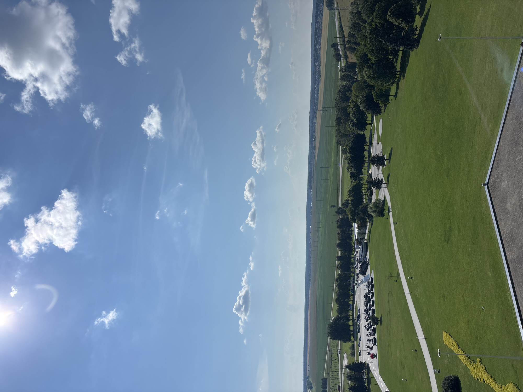 The image captures an expansive outdoor scene under a bright, blue sky dotted with scattered white clouds. The sun, high and slightly to the left, casts a strong light, creating a halo effect. In the foreground, the edge of a dark, textured roof suggests an elevated viewpoint. Below, a vast, meticulously maintained green lawn stretches out, traversed by a winding paved walkway. Along the left edge of the lawn, a vibrant yellow-green hedge forms an angular shape.
Further into the midground, a parking lot is visible, filled with numerous cars. Adjacent to the parking area are several trees, some forming a dense line, others standing individually. Beyond the parking lot and trees, a transition occurs to cultivated fields, appearing as broad swathes of green and lighter brown, indicative of agricultural land. In the distance, a line of trees marks the horizon, with a hint of scattered buildings and infrastructure such as power lines. The overall environment suggests a park or recreational area adjacent to agricultural land, likely in a suburban or rural setting within the Ozyaritska-Slabadski rural council, Belarus. No people are clearly discernible in the image, and no specific actions or interactions are depicted.
