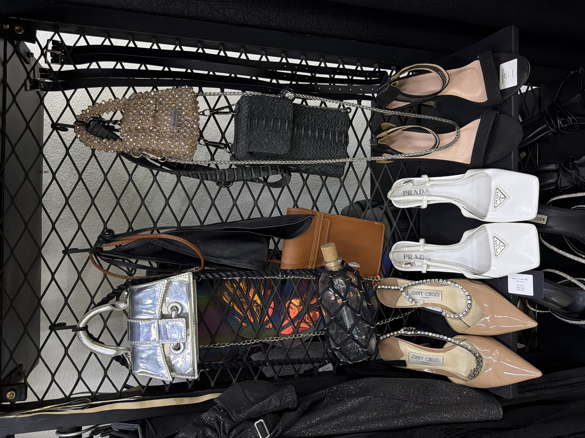 The image shows a retail display of women's luxury accessories, primarily handbags and shoes, hanging on a black metal grate shelf. The items are arranged in a visually appealing manner, with handbags displayed at the top and shoes at the bottom. The overall aesthetic is one of a high-end boutique, with a focus on designer brands. There are no people present in the image. The background is a plain white wall, suggesting an indoor retail environment. The lighting is bright and even, highlighting the details of the products. Based on the merchandise and presentation, this scene is likely from a boutique located in North Kuta, Indonesia, which is known for its luxury shopping destinations.