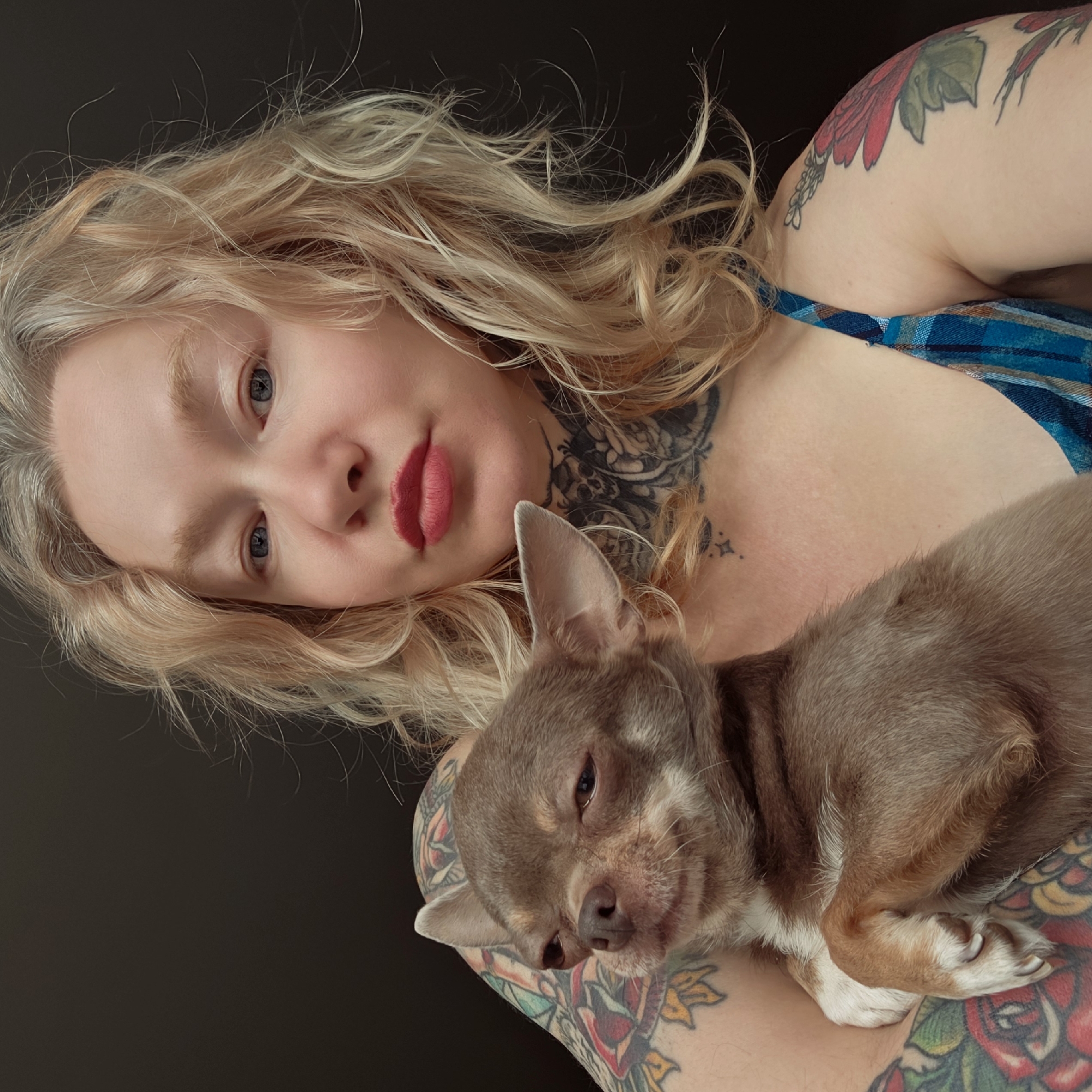 A woman with blonde hair and tattoos is holding a brown chihuahua. The woman has fair skin, blue eyes, and red lipstick. She is wearing a blue plaid top. The chihuahua is lying on her arm, appearing relaxed or sleepy. The background is a dark, solid color. The location is not discernible from the image.