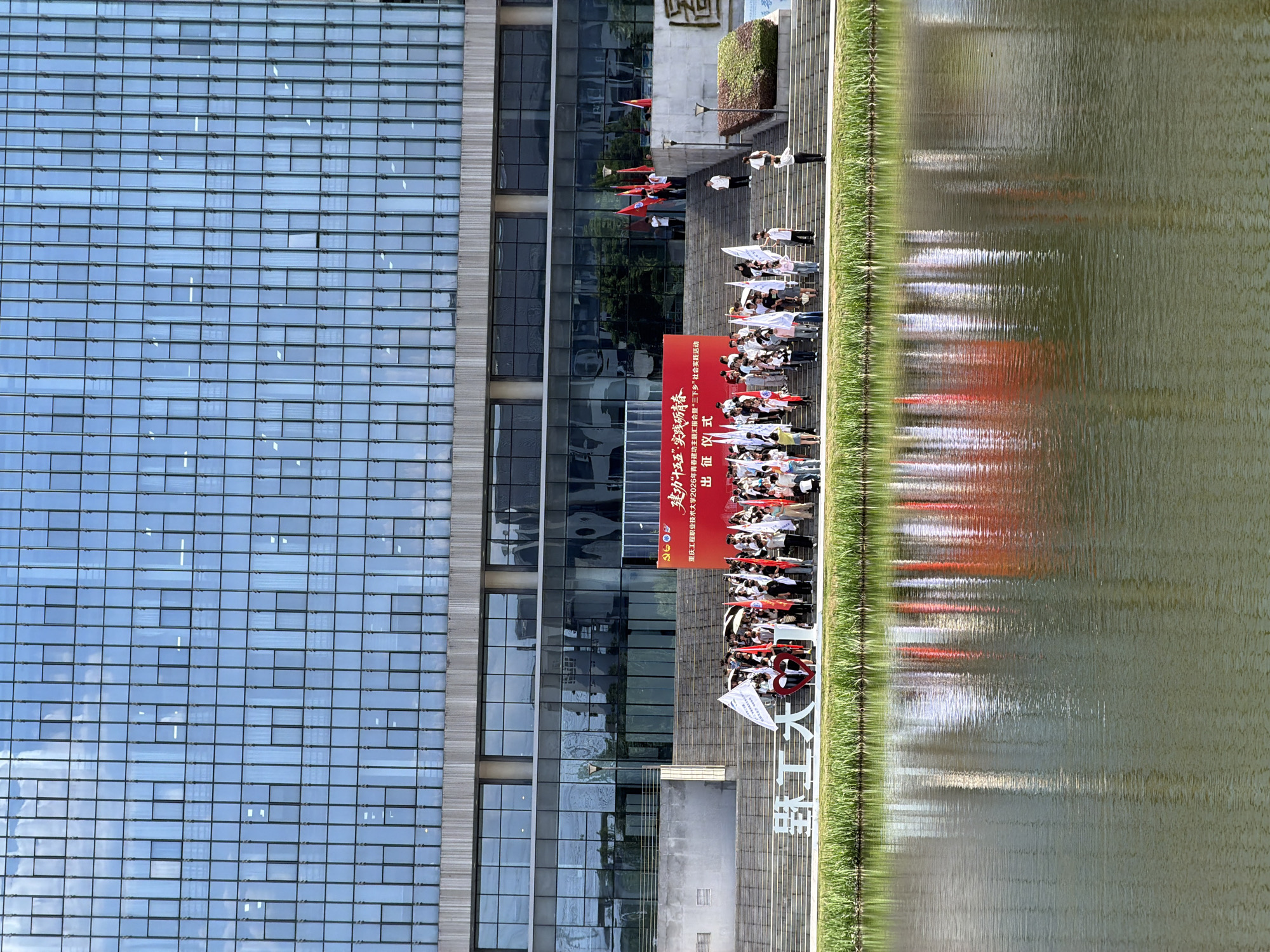 This is a wide shot of a formal gathering in front of a modern building with a glass facade, likely a university or office complex, at dusk. A large body of water, possibly a river or lake, occupies the foreground, reflecting the building and the event. A group of people are gathered on a set of wide, concrete stairs leading up to the building. They are holding numerous red and white flags, and some appear to be holding umbrellas, suggesting the event might be a ceremony or a rally. A large red banner with Chinese text is displayed on the stairs. In front of the stairs, a large white sign with Chinese characters, possibly spelling out the name of the location or institution, is visible. The overall atmosphere appears to be one of an organized event with a sense of officialdom. The location is identified as Jiangjin, China, based on the context provided in the prompt.