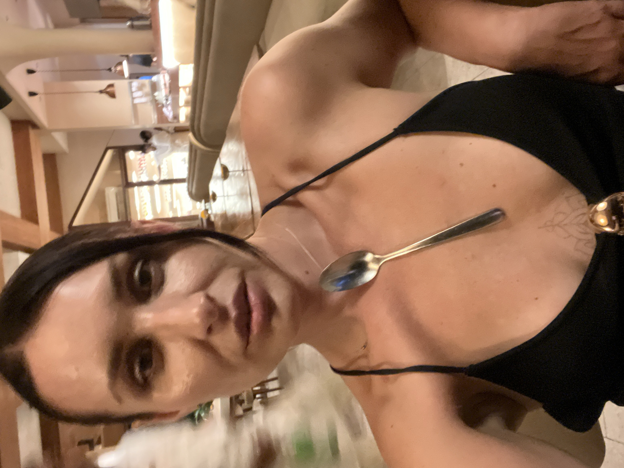 A selfie shows a woman with brown hair and eyes, wearing a black top with thin straps. A silver spoon is balanced on her chest. The background appears to be a restaurant interior with light fixtures and wooden structures. While the image suggests an indoor location, it does not provide enough specific detail to definitively determine if the location is in Kuta Selatan, Indonesia.
