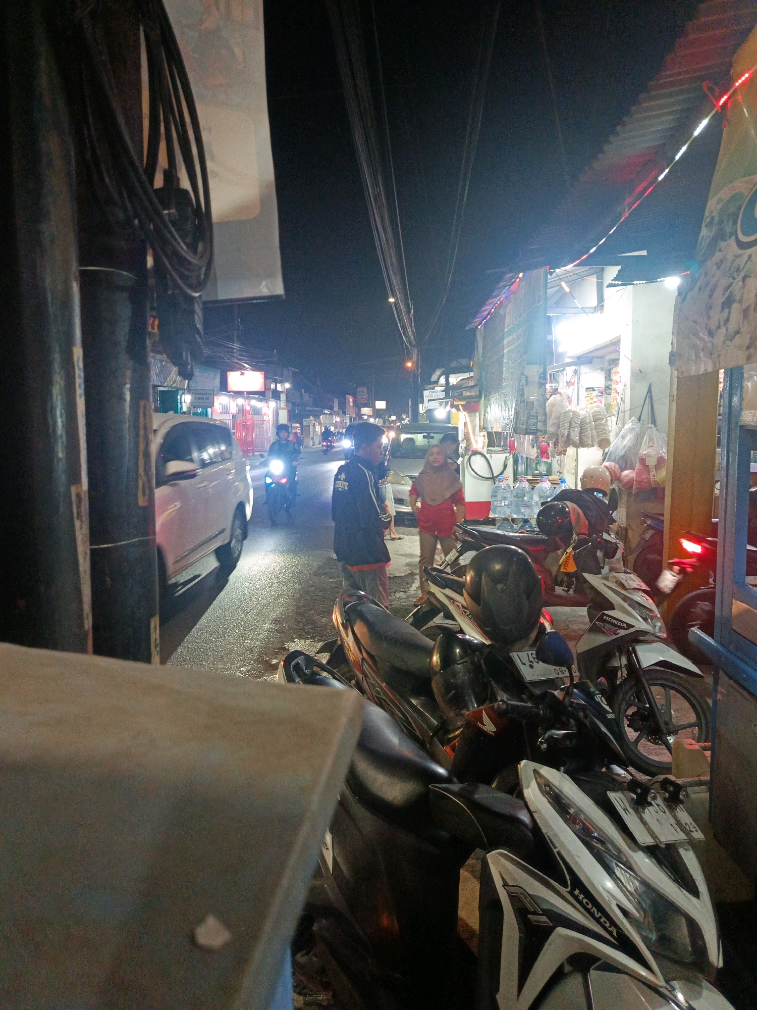 In Sedati Agung, Indonesia, at night, a street scene unfolds under artificial light. A white van is parked on the wet asphalt road, its headlights illuminating the area. A person on a motorcycle with its headlight on rides past the van. In the foreground, several motorcycles are parked, some with helmets on them. A person in a red top stands near a stall displaying bottled water. Another person, wearing a dark jacket with red accents, is also present. The street is lined with buildings, some of which have illuminated signs and decorative lights. Wires are strung across the overhead view, typical of urban infrastructure. The atmosphere suggests a busy commercial area at night.