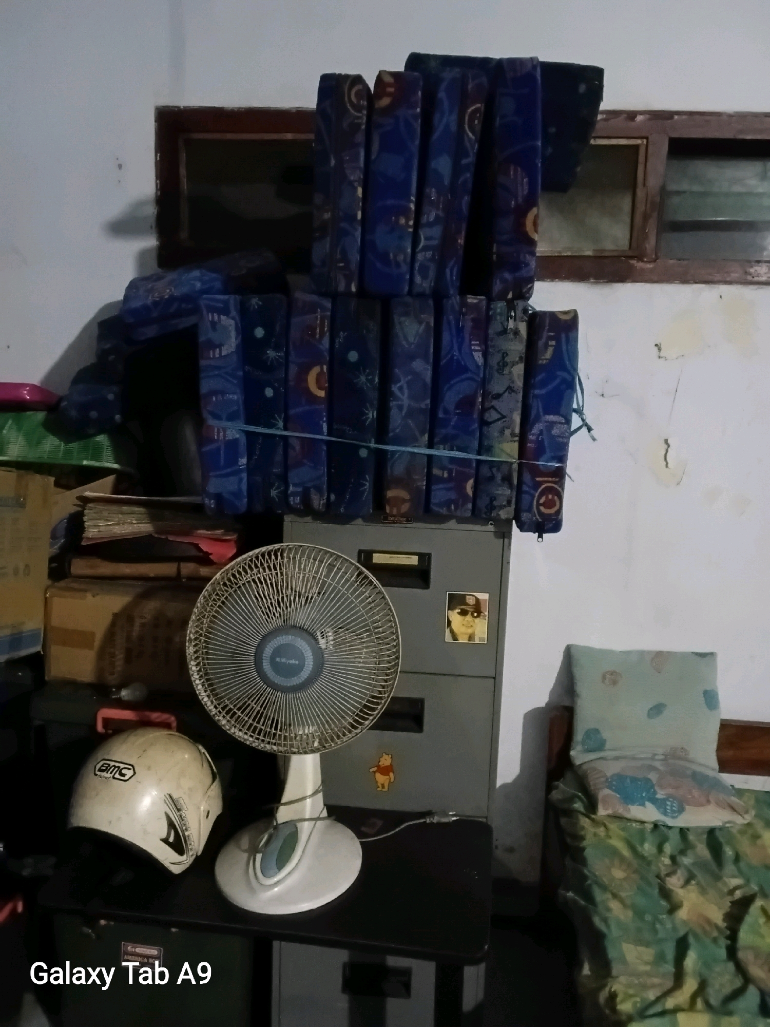 The image captures an indoor scene, likely a bedroom or storage area, in Kelutan, Indonesia. A white BMC brand helmet rests on a black surface to the left of a white electric fan. Behind the fan, a grey filing cabinet is visible with a small sticker of a person in sunglasses and a hat on its front. To the right of the cabinet, a bed is partially visible, covered with a green patterned duvet and adorned with two pillows, one light blue with a faint pattern and another with blue and pink motifs. A collection of blue, patterned upholstery pieces are stacked vertically behind the filing cabinet, appearing to be seats or cushions. Cardboard boxes and stacks of paper are arranged on the left side of the image, beneath the stacked upholstery. The room has a light-colored wall with visible imperfections and damage. A wooden-framed window with glass panes is situated in the background above the stacked upholstery. The overall impression is of a cluttered, lived-in space.