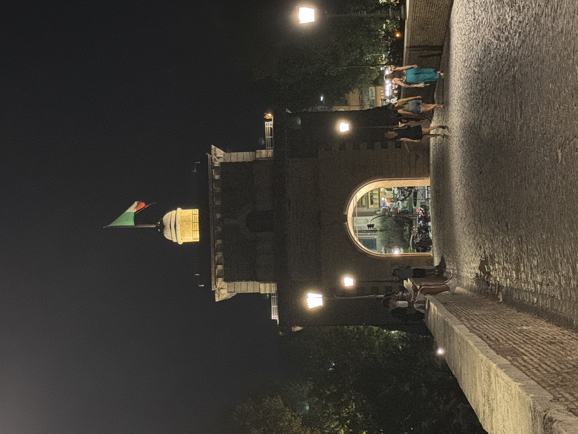 The image depicts the Ponte Sant'Angelo in Rome, Italy, at night. An illuminated archway, part of a historic structure, forms the central focus. An Italian flag flutters from atop a turret above the archway. The bridge is paved with cobblestones, and stone walls line the sides. Several people are present on the bridge; some are walking towards the archway, while others are seated on the wall. Streetlights cast a warm glow on the scene, illuminating the pathway and surrounding trees. The atmosphere is that of a lively evening in a historical urban setting.