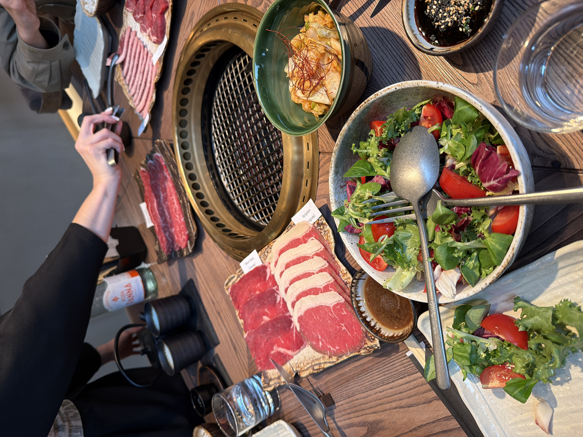 The image captures a close-up, overhead view of a wooden table set for a meal, likely a Korean BBQ or a similar dining experience. A built-in grill is centrally positioned, its grates clean and ready for use. Around the grill, various food items and utensils are arranged. Slices of raw red meat, possibly beef, are laid out on platters, some in thin, uniform slices and others in thicker cuts. A bowl of vibrant green salad, mixed with red tomatoes and other leafy greens, is prominent in the foreground. Adjacent to the salad are small dishes containing sauces, one appearing to be a dark dipping sauce with sesame seeds, and another a lighter brown sauce. A bowl of kimchi, a traditional Korean fermented cabbage dish, is also visible, garnished with red chili threads.
On the left side of the table, a glass of water is partially visible, along with two dark ceramic cups, possibly for beverages. A bottle of San Pellegrino water with its distinctive label is placed behind these cups. In the upper right portion of the image, a person's hands are visible, one holding a pair of tongs, suggesting active engagement in the cooking process. The background is slightly blurred, but it appears to be an indoor setting with a muted color palette.
There is no specific visual information in the image to suggest the location is Riga, Latvia.
