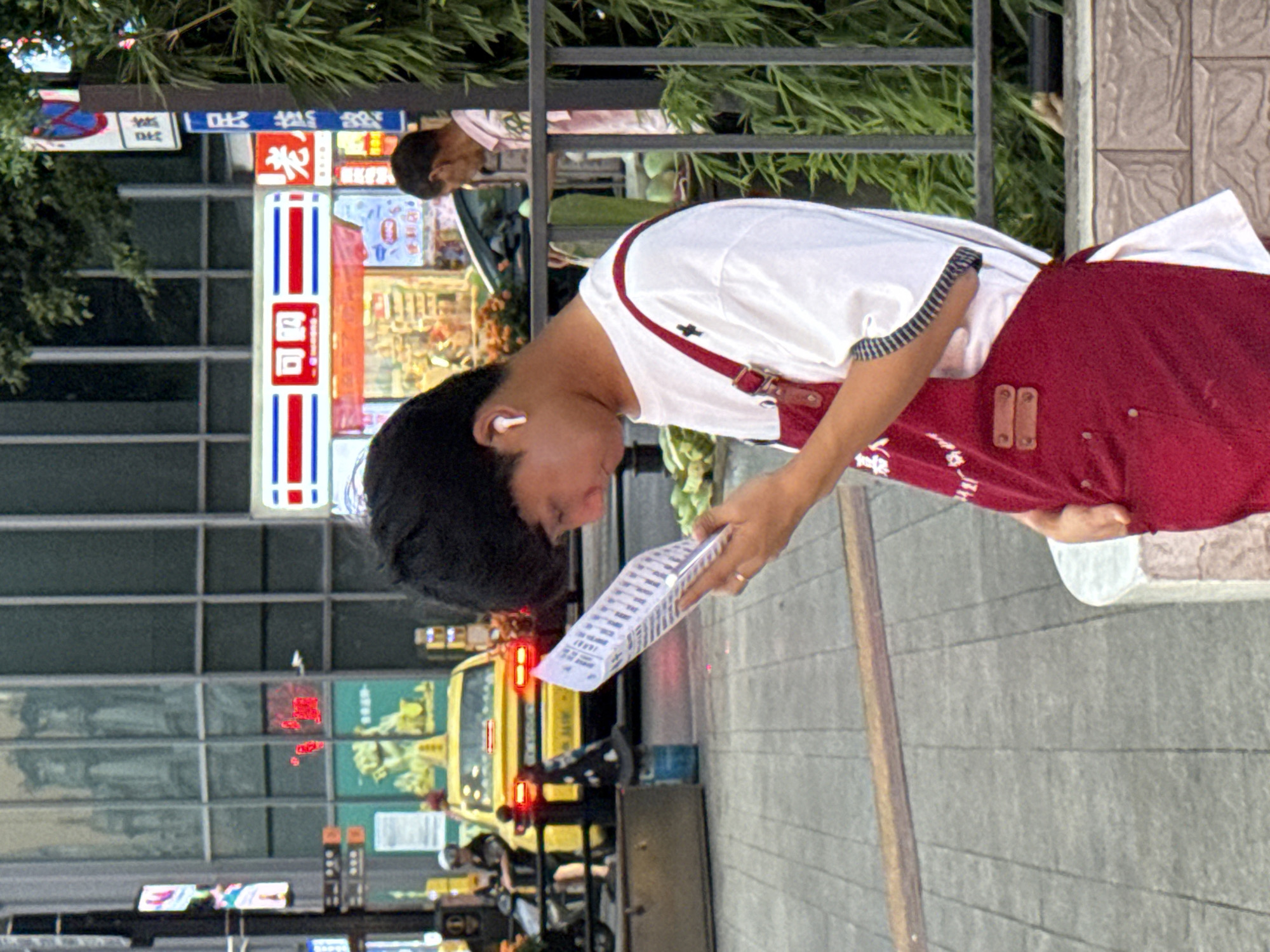 A young Asian male is standing on a sidewalk in a city, holding a white paper and looking down at it. He is wearing a white t-shirt with black trim on the sleeves and a red apron over it. The apron has two metal buttons on the bib and black straps that go over his shoulders. He has short black hair and is wearing white earbuds. Behind him, a yellow taxi is stopped at a traffic light. In the background, there are buildings, a convenience store with a red and white sign, and a lamppost with signs. To the right of the man, there is a metal fence and some greenery, including bamboo. The street is paved with gray tiles. The overall scene appears to be a street in a busy urban area.