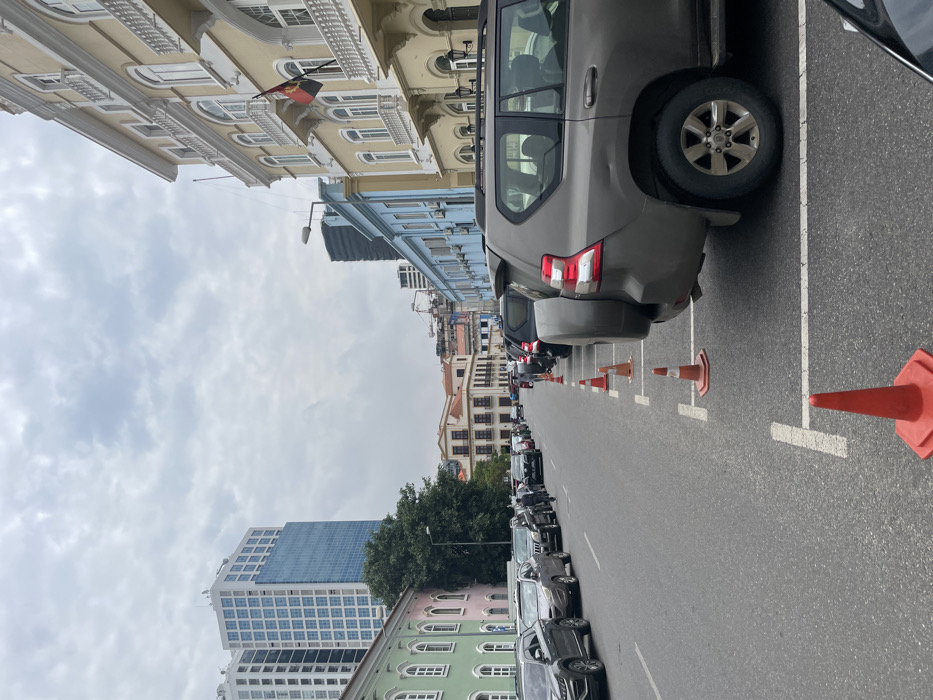 The image captures a street scene in Luanda, Angola, under an overcast sky during the daytime. The perspective is down a wide asphalt road marked with white parking lines. A row of parked cars, predominantly SUVs, lines the right side of the street, delineated by orange traffic cones. The closest vehicle is a gray SUV with a spare tire mounted on its rear.
On the left, a varied architectural landscape unfolds. A prominent multi-story building, painted light yellow with ornate white window frames and balconies, suggests colonial-era design. From this building, a distinctive Angolan flag, red and black with a yellow emblem (machete, cogwheel, star), is visibly hanging. Further along the street, other buildings, including a light blue structure and several in pastel shades, line the road. In the background, modern high-rise buildings rise above the older structures, illustrating the urban development of the city. A small cluster of green trees is also visible. The sky is uniformly gray, indicating a cloudy day. No people or legible text are visible within the frame.