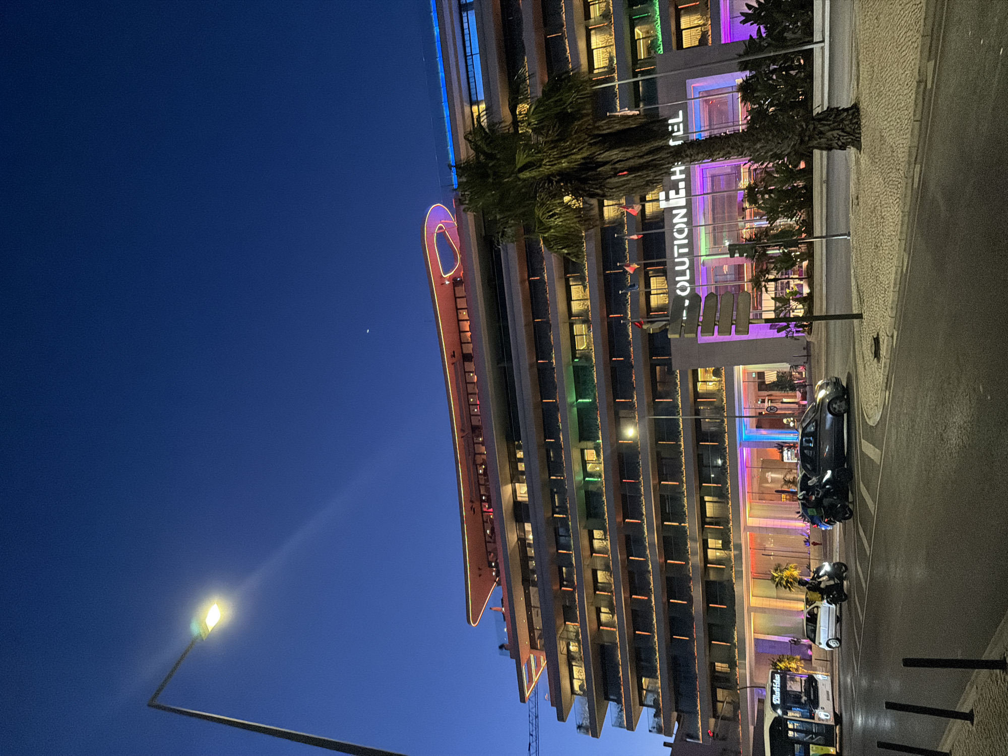 The image depicts a modern, multi-story hotel named "E.HOTEL" located in Cascais, Portugal, at dusk. The hotel features a distinctive curved architectural element at the top, illuminated by colorful lights. Each floor of the building has multiple windows, with some showing warm interior lighting. The facade is adorned with horizontal bands of colored lights, predominantly in shades of green, orange, and purple, which are reflected on the street.
In front of the hotel, a palm tree stands tall. Several flags are displayed near the entrance. A street with crosswalk markings runs in the foreground. A white bus with "Dila Eles" written on its destination board is visible on the left. A white car and two motorcycles, one ridden by a person with a delivery bag, are also present on the street, appearing to be at an intersection. A street lamp with a lit bulb illuminates the scene. The sky is a deep blue, indicating it is twilight.