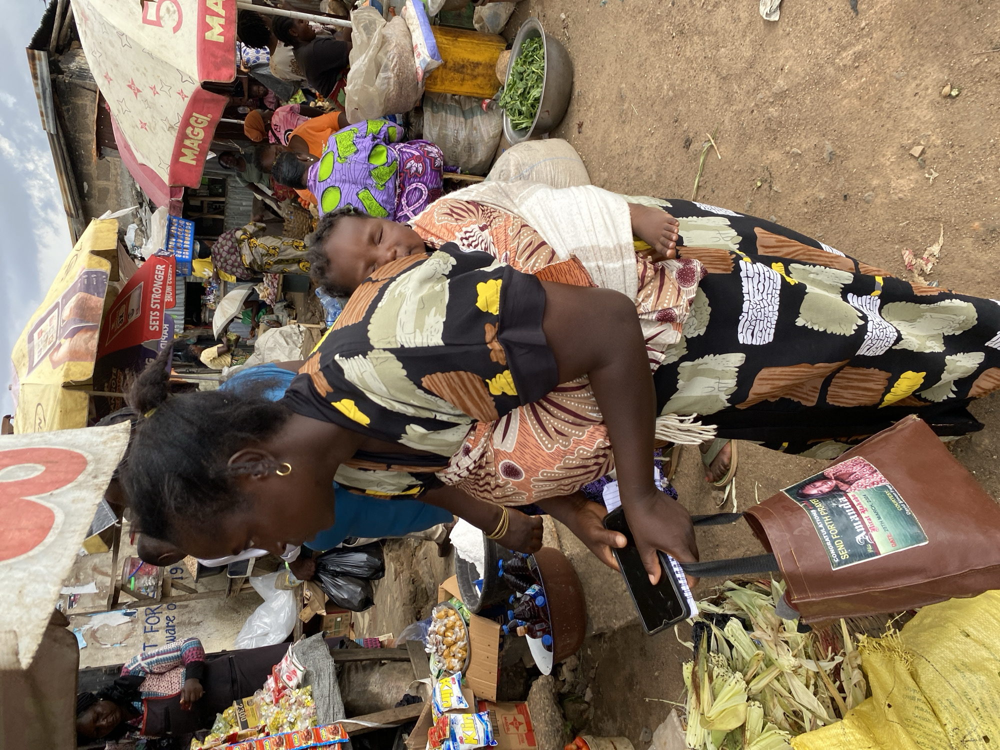 A Black woman in Bukuru, Nigeria, carries a baby on her back. The woman is wearing a black and colorful patterned dress and a gold hoop earring. She holds a smartphone and a notebook in her left hand and a brown tote bag with a picture of a woman and text in her right hand. The baby, also Black, is wrapped in a light-colored cloth. In the background, an outdoor market is visible with various vendors, stalls, produce, and umbrellas. The ground is dirt.
