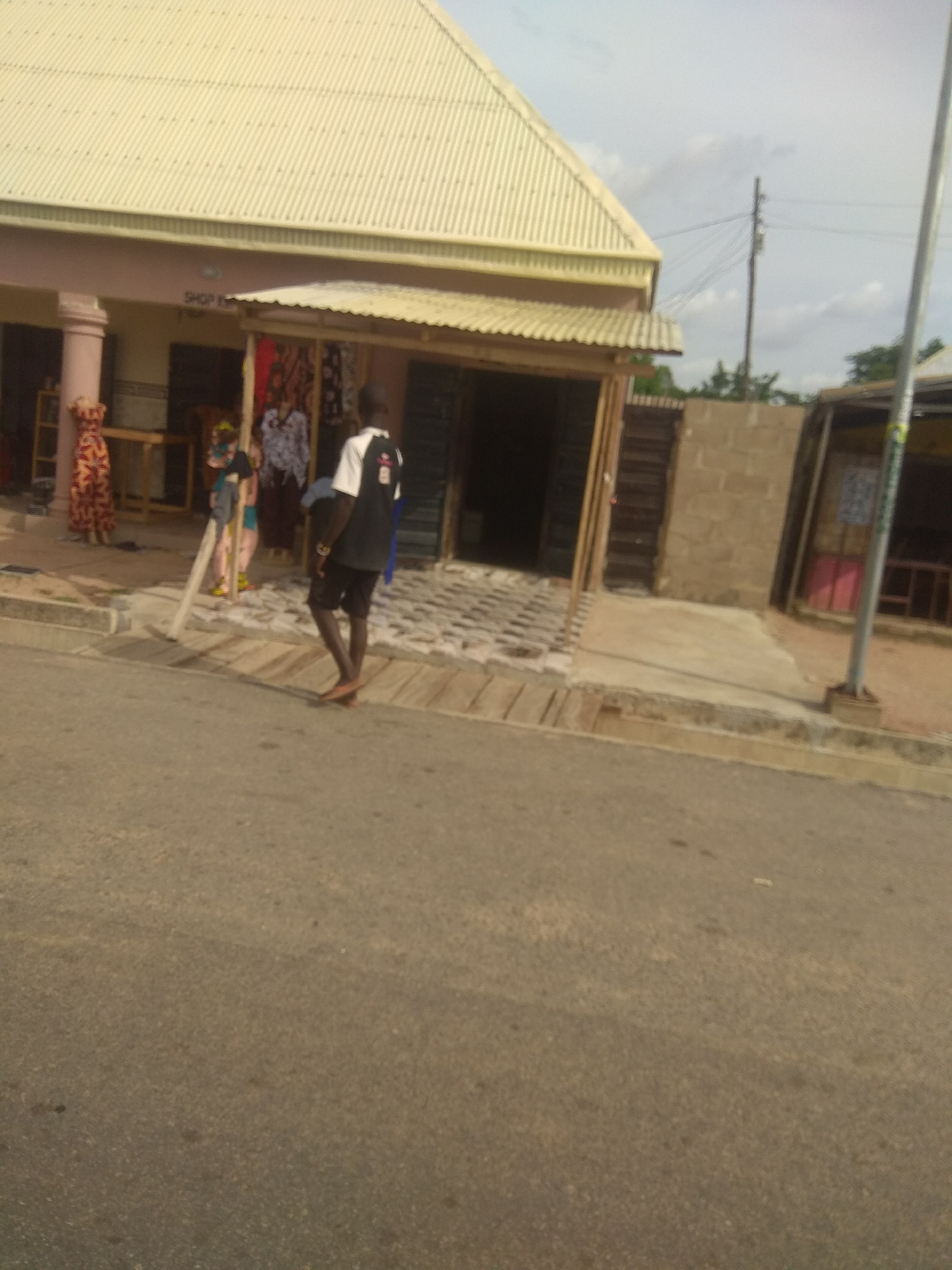 The image depicts a street scene in Jalingo, Nigeria, with a row of small shops on the right side. In the foreground, a man wearing a black and white t-shirt and dark shorts walks away from the camera, with his left foot extended. He is wearing light-colored sandals. Behind him, a woman in a colorful, patterned dress stands on the left side of a storefront. Another person, possibly a woman, is visible behind the first woman, wearing a patterned top and blue pants. To the right of the first woman, a man wearing a patterned shirt and dark pants stands near the entrance of a shop. The shops have corrugated metal roofs and open fronts displaying merchandise. The ground in front of the shops is paved with interlocking stones. A paved road runs in front of the shops. In the background, a utility pole with wires is visible, and trees can be seen beyond the buildings. The sky is overcast. The overall impression is of a busy, commercial area.