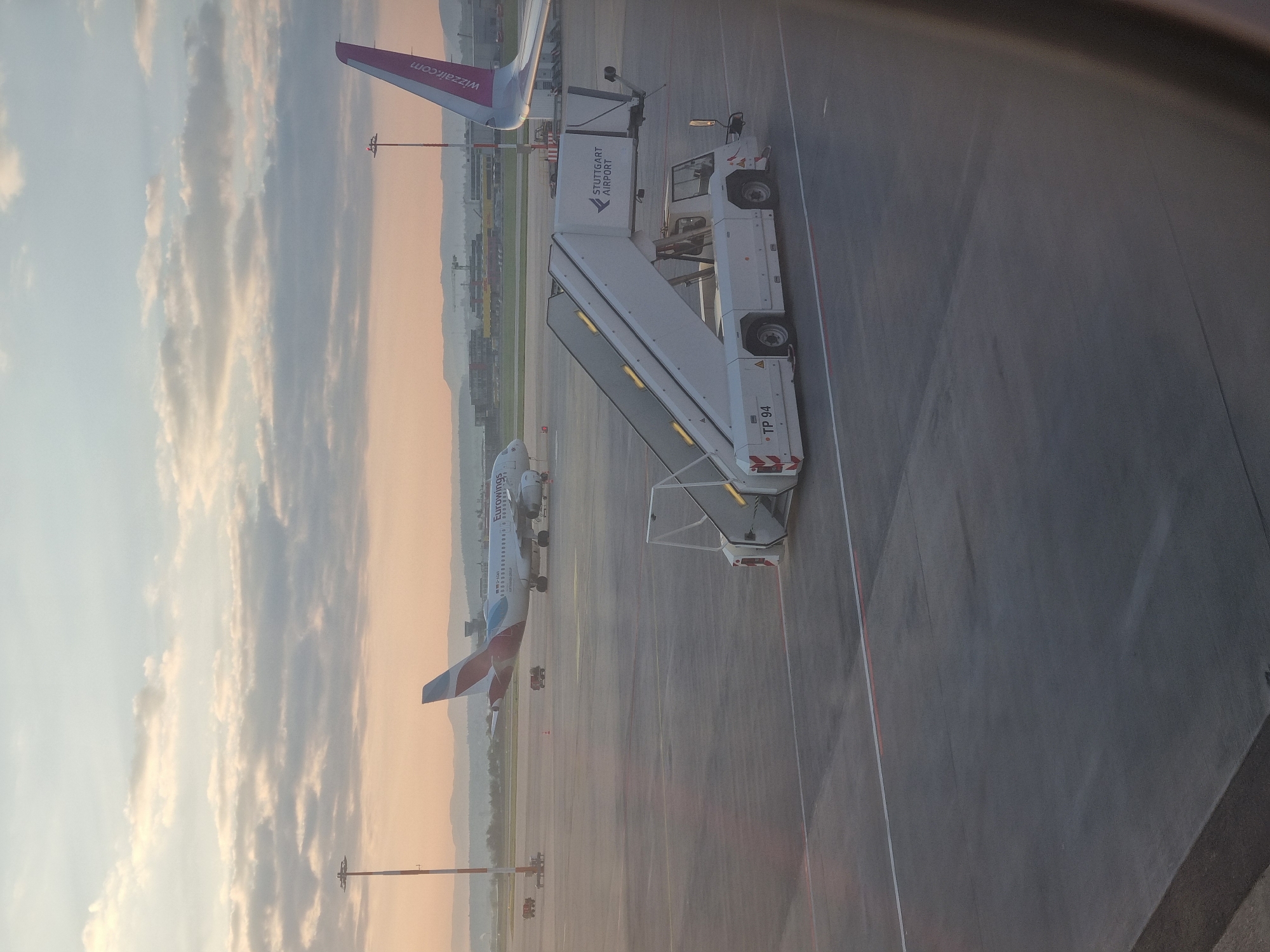 The image shows a view from an aircraft window at Stuttgart Airport (STR) in Echterdingen, Germany. Two airplanes are visible on the tarmac: a Eurowings Airbus A320 and a Wizz Air airplane. A passenger boarding bridge with "Stuttgart Airport" branding is positioned on the tarmac near the window. The sky has a mix of blue and pink hues, suggesting either sunrise or sunset. Ground equipment and infrastructure are visible in the background, including lighting poles and other airport vehicles. The ground is concrete with painted lines.