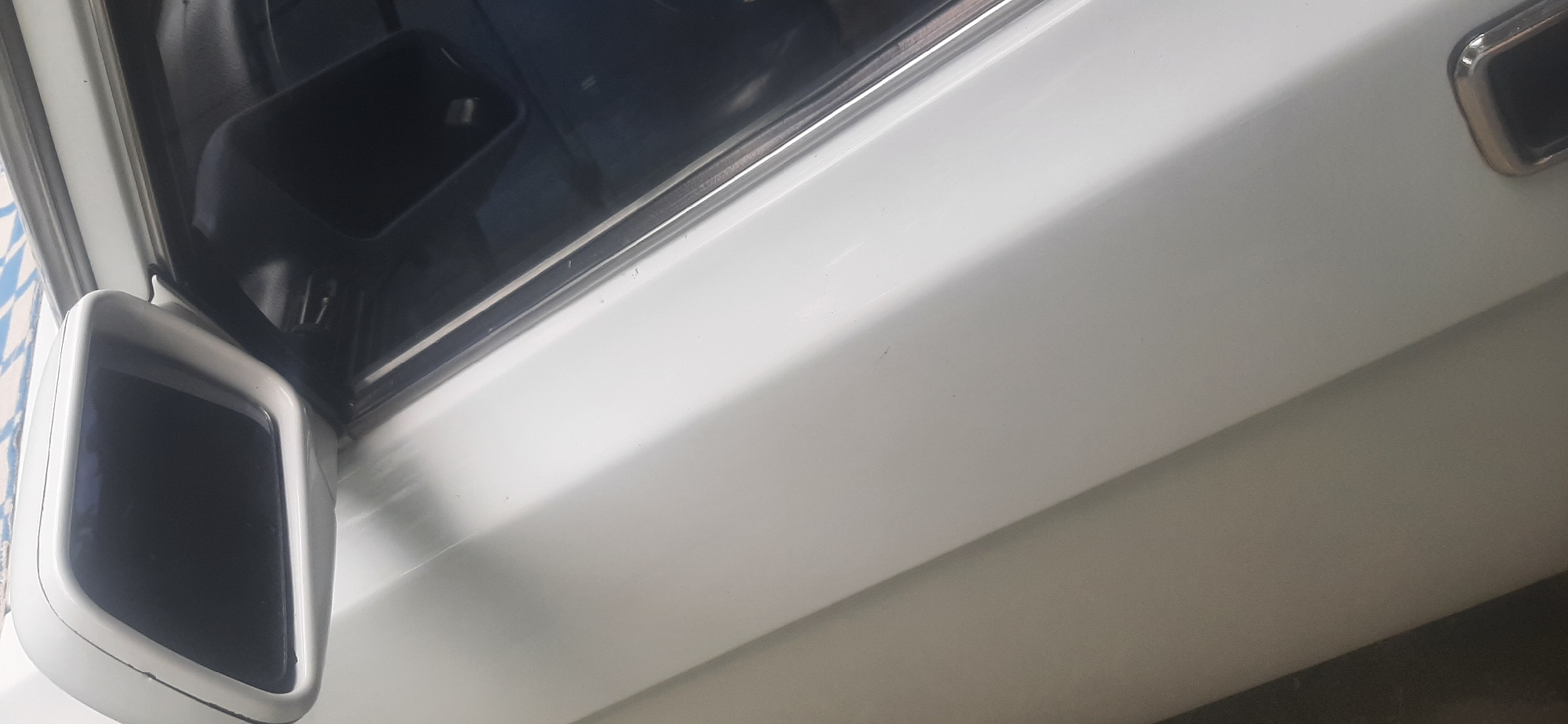 The image displays a portion of a white car's exterior, focusing on the driver's side door and side mirror. The side mirror is mounted on the door and has a black reflective surface. A window frame with a chrome accent is visible along the door edge. The car appears to be in a clean, well-maintained state. The background is out of focus, but suggests an outdoor setting, possibly a street or a parking area in Yessentukskaya, Russia. There are no people or any specific landmarks visible that would further pinpoint the location within the city.