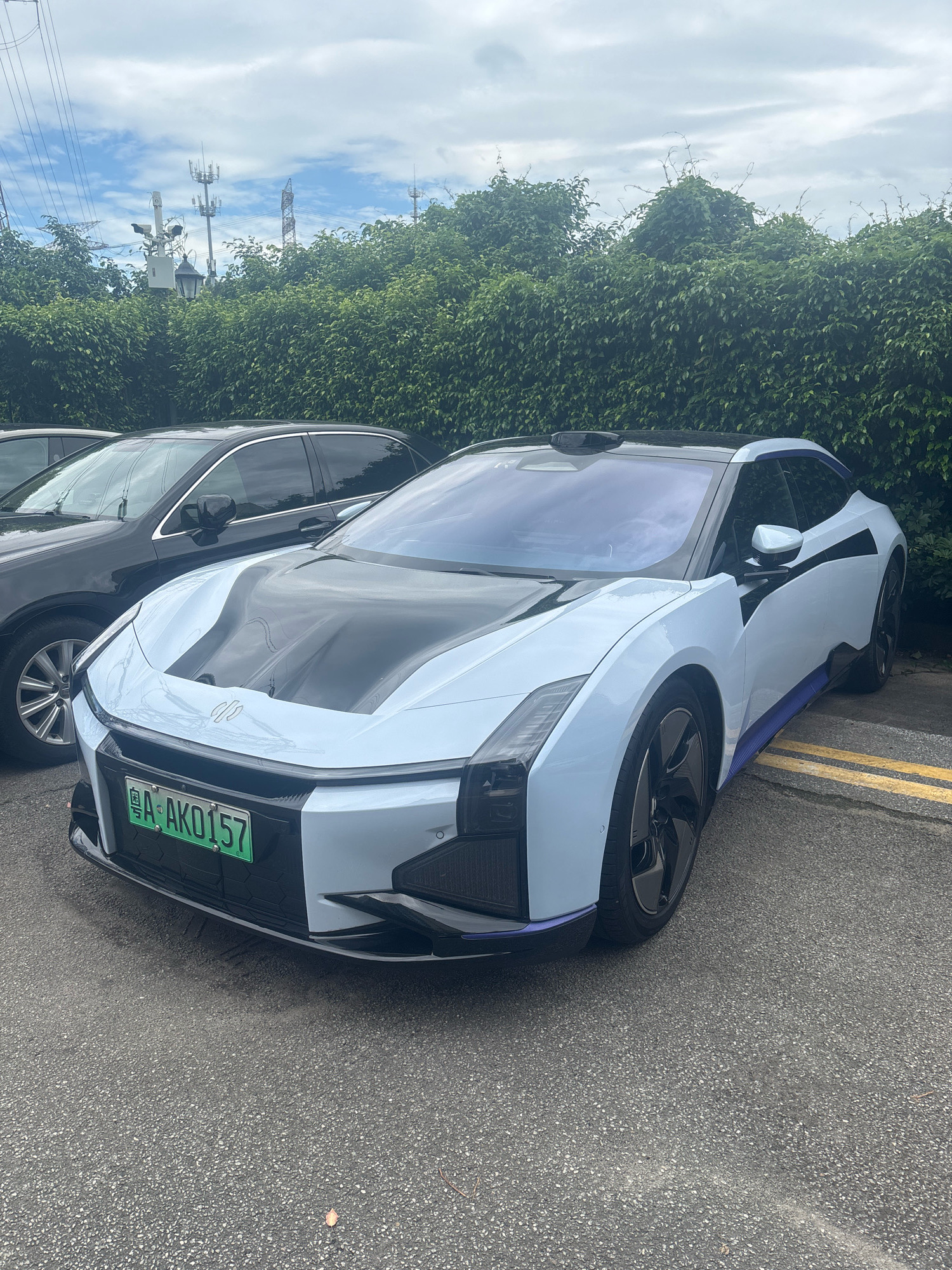 A light blue electric car with a black hood is parked on an asphalt surface in front of a dense green hedge. The car has a prominent license plate that reads "A AK0157". Behind the car and hedge, there are utility poles and cell towers visible against a cloudy sky. The car's design is futuristic, with sharp lines and angular features. It appears to be a bright, overcast day. The car is positioned on the right side of the frame, with another dark-colored vehicle partially visible to its left. Yellow lines marking a parking space are visible on the asphalt in front of the car.
