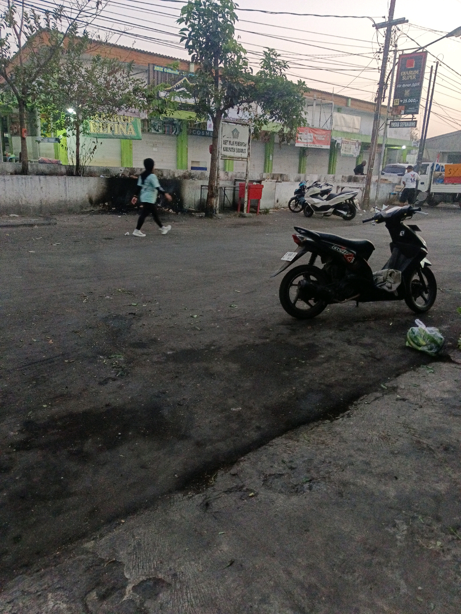 The image shows an outdoor scene in the city of Wonocolo, Indonesia, at what appears to be dusk. In the foreground, a dark-colored scooter is parked on a paved surface. To the left, a person wearing a light blue hijab and dark pants walks away from the camera. Behind them, a low concrete wall is visible, with trash and debris piled against it. Beyond the wall, several buildings with green and white facades are lined up, some with closed shutters. Trees with sparse foliage are interspersed along the front of the buildings. A tall utility pole with numerous wires stands on the right side of the image. In the background, there are signs on the buildings, including one that reads "DIARUM SUPER" and another mentioning "PASAR SEPANJANG". Several other motorcycles are parked in front of the buildings. A white truck is also visible in the background to the right, with a person wearing a white shirt and black pants standing near it. The ground surface is predominantly dark asphalt with patches of lighter concrete and scattered debris. A plastic bag containing green items is on the ground near the scooter in the foreground.
