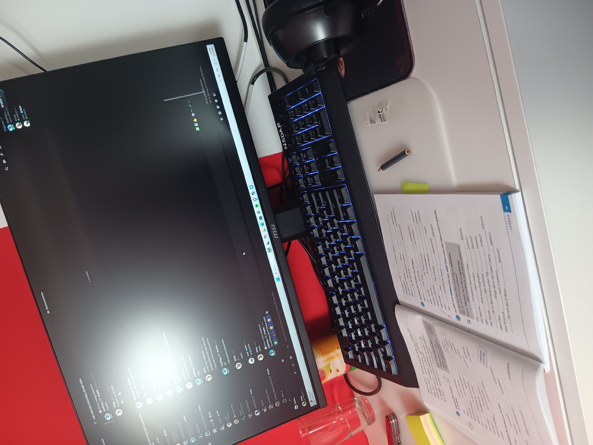 The image displays a desk setup in Sofia, Bulgaria, captured from an elevated, angled perspective. The scene centers around a computer monitor, a mechanical keyboard, and an open textbook, indicating a study or work environment.
A black MSI brand monitor occupies the left side, displaying a desktop interface with an "Activate Windows" watermark. The Discord application is prominently featured, showing active chat messages in Bulgarian, English, and some German. Notable text includes "FREI UND DEUTSCH WOLLEN WIR SEIN", discussions about homework ("sq proq domashnoto"), gaming ("AFK v edna igra"), and friendly banter. Usernames such as "BobPlays YT", "dang", "meech", and "meefisch" are visible. The system clock shows "5:27 PM" on "4/27/2026", while some Discord messages are dated "21. April 2024".
Adjacent to the monitor is a black mechanical keyboard with blue backlighting, notably featuring "NSDAP" branding on its left side. Behind the keyboard, a pair of black headphones rest on a mousepad, which bears a distinctive Razer logo.
On the white desk surface to the right, an open German language textbook is visible. Page 19 contains exercises on the "Perfekt" tense, with instructions like "Ergänze die Verben im Perfekt" (Complete the verbs in the Perfect tense) and "Übersetze die Sätze und vergleiche" (Translate the sentences and compare). Various German verbs and sentences are presented for language practice. A yellow highlighter and a pencil are placed on the book. Other items on the desk include a red pocket knife, a clear glass, and an orange container. A red surface is partially visible beneath the monitor and keyboard.
The overall scene depicts a person engaged in language learning, possibly German, while also interacting with online communities via Discord.