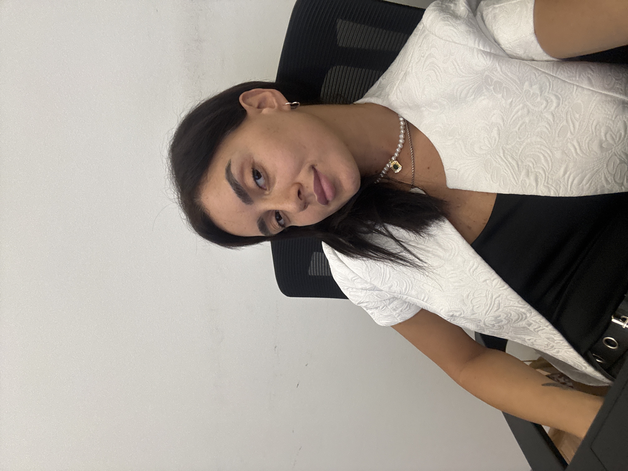 A woman is sitting in a chair in front of a white wall, looking at the camera. She is wearing a white jacket with a textured pattern over a black top. She has dark hair and is wearing a pearl necklace with a pendant. The scene appears to be an indoor setting, possibly an office or a studio.