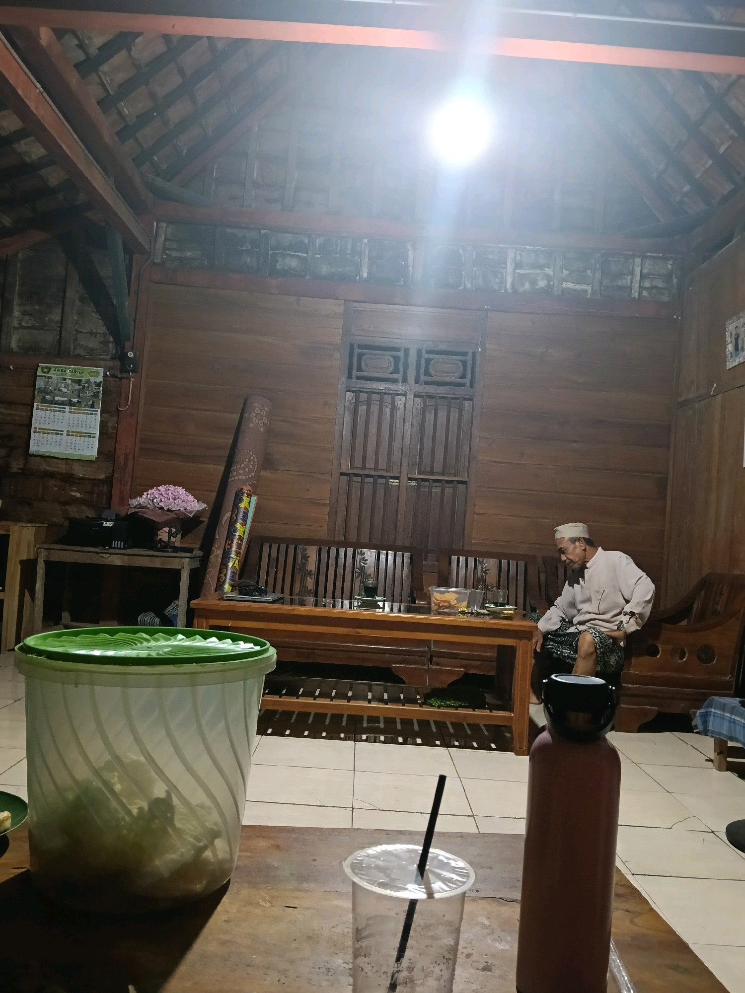 The image depicts an indoor scene inside a wooden structure, likely a home, in Galuk, Indonesia. The room has a high, sloped ceiling with exposed wooden beams and rafters, covered with clay tiles. A bright overhead light fixture illuminates the space. The walls are paneled with dark wood. In the foreground, a wooden table is visible, with a large, translucent green plastic container filled with what appears to be food or ice on the left. To the right of this container is a clear plastic cup with a black straw. Further to the right is a tall, reddish-brown thermos bottle with a black lid. In the mid-ground, a long, wooden bench with cushioned seating is positioned against the back wall. A man, appearing to be elderly, is seated on this bench, wearing a light-colored long-sleeved shirt, a patterned sarong, and a white peci cap. He is looking down and to his left. In front of the bench is a lower, wider wooden table with items on its surface, including what looks like a plastic container of snacks and a cup. A rolled-up rug with a decorative pattern leans against the wall behind the bench. To the left of the bench, a smaller wooden table holds a bouquet of pink flowers and what appears to be electronic equipment. A calendar with images and text, likely advertising, hangs on the wooden wall to the left of the bench. The floor is tiled with square, light-colored tiles. The overall impression is a casual, domestic setting.