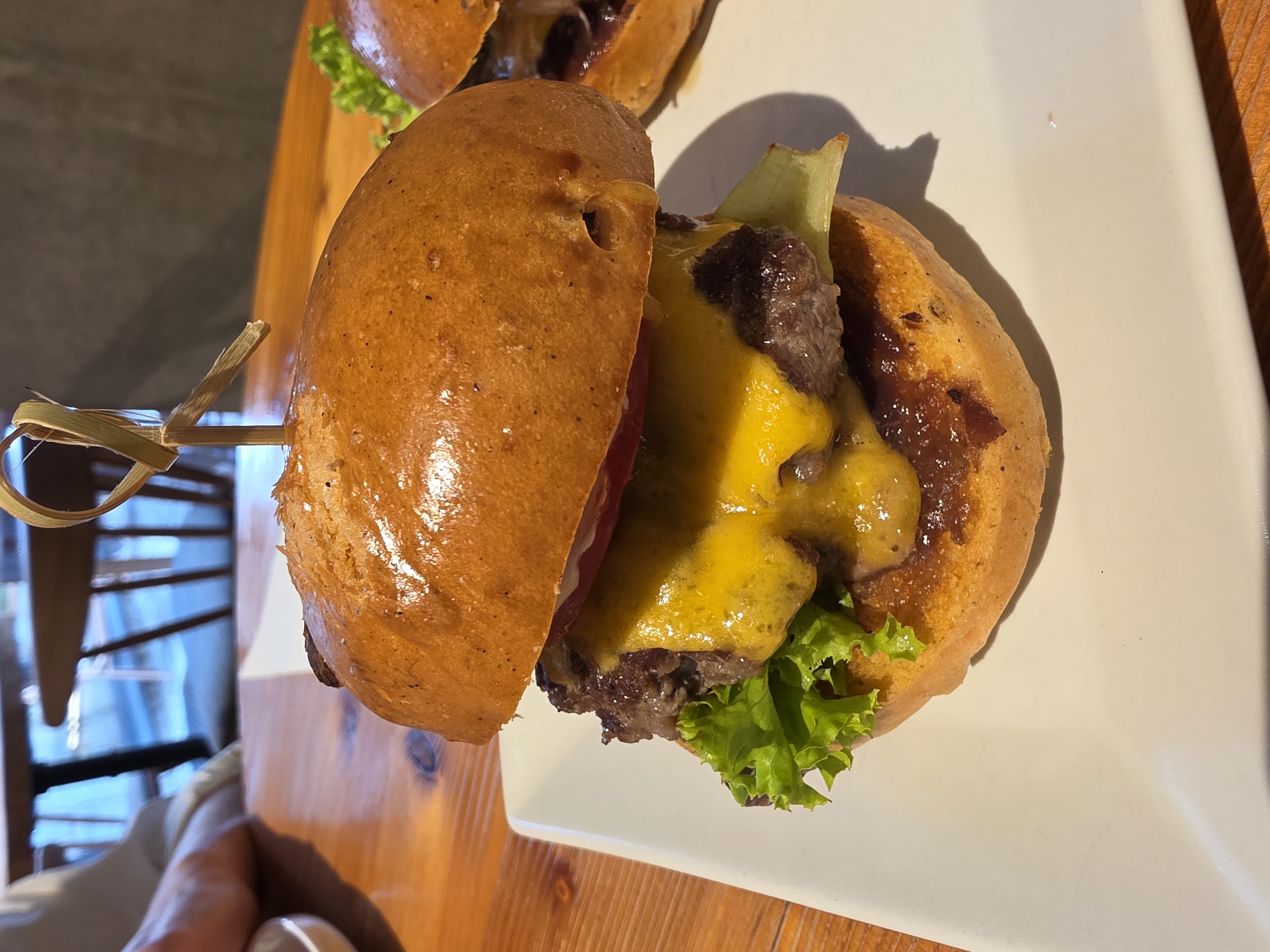 A close-up, eye-level shot captures a gourmet cheeseburger presented on a white rectangular plate. The burger, centered in the frame, features a golden-brown toasted bun, a thick, juicy beef patty, melted bright yellow cheese dripping down the sides, and fresh green lettuce. A skewer made of natural fibers pierces the top bun, its ends fanning out. In the background, a wooden table is visible, with out-of-focus chairs and a person's arm resting on the table. The scene appears to be set in a restaurant.