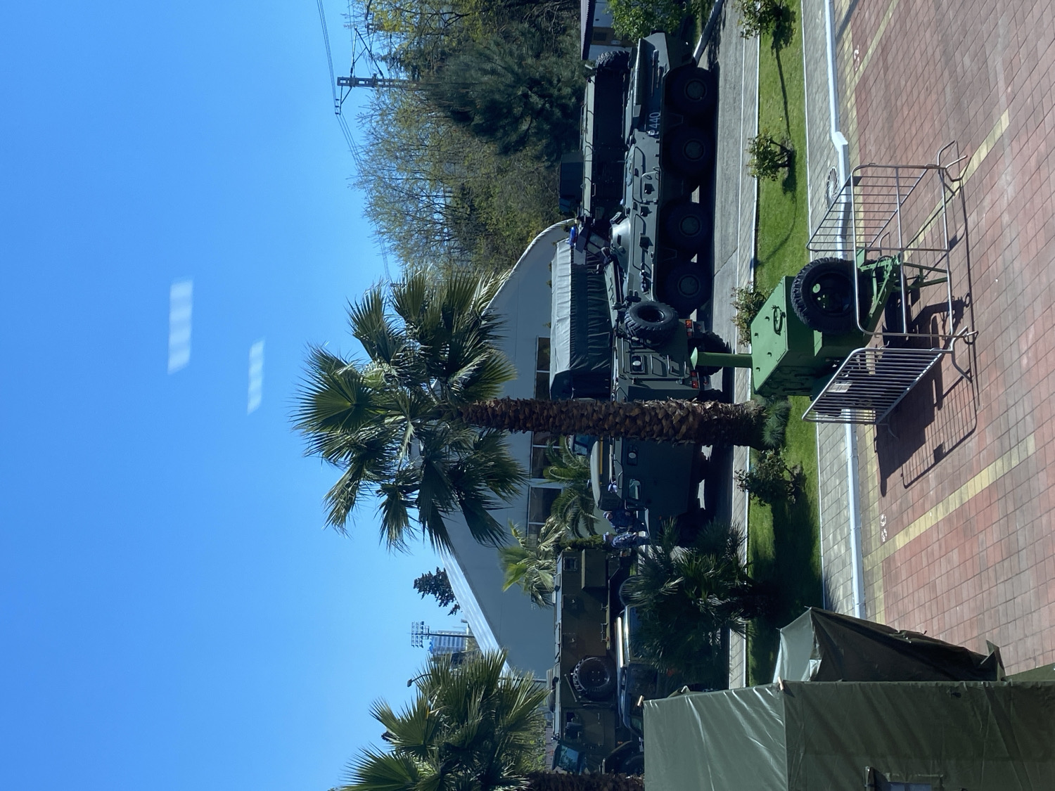 The image displays an outdoor scene featuring a collection of military equipment under a bright, clear blue sky. Several olive-green military vehicles are arranged in a line on a paved surface, which consists of reddish-brown interlocking pavers with white demarcation lines. These vehicles include what appear to be armored personnel carriers or infantry fighting vehicles and military transport trucks. One of the vehicles prominently displays the alphanumeric designation "E 440" on its side.
Adjacent to the vehicles, a green military trailer, equipped with a visible spare tire, is parked near a metal barrier or wheeled cart. In the foreground, two large, olive-green military tents are set up on a patch of grass. The landscape is dotted with several tall palm trees, indicating a warm or subtropical climate. In the background, deciduous trees and utility poles with power lines are visible against the sky.
The scene is illuminated by natural sunlight, casting distinct shadows, suggesting it is daytime. The weather appears clear and sunny. No people are visible in the image. The arrangement of the vehicles and tents, along with the absence of active personnel, suggests a static display, exhibition, or organized temporary encampment of military assets.